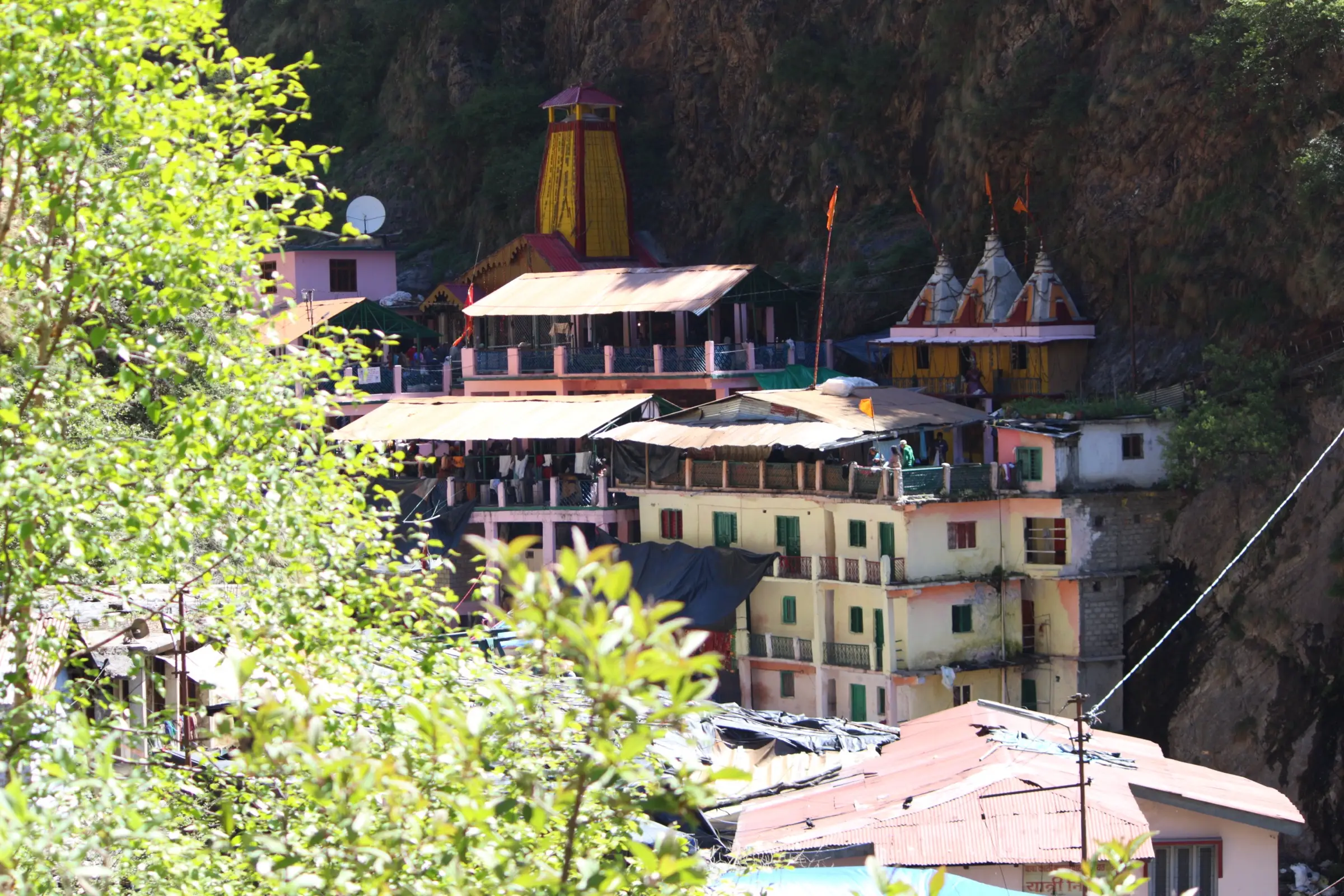 The yellow spire of the Yamunotri temple and pilgrim buildings against a dark cliff, seen through green leaves
