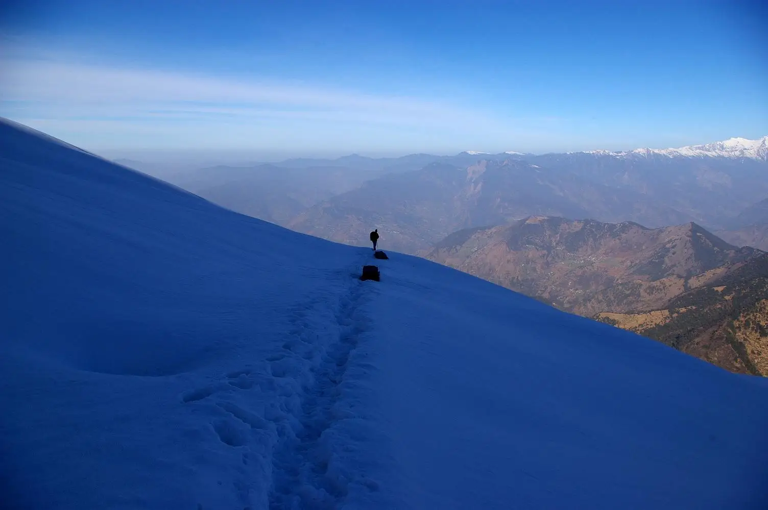 A lone trekker on a snow-covered ridge path towards Tungnath with brown hills and distant snow peaks below