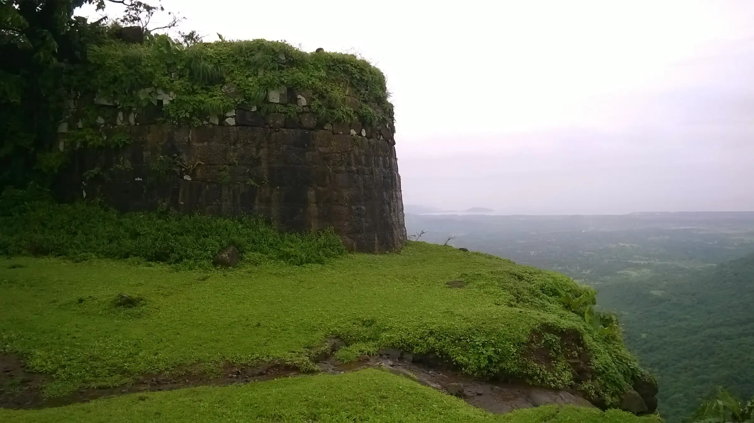 Round stone south bastion of Sagargad fort on a green grassy edge with the misty plain below