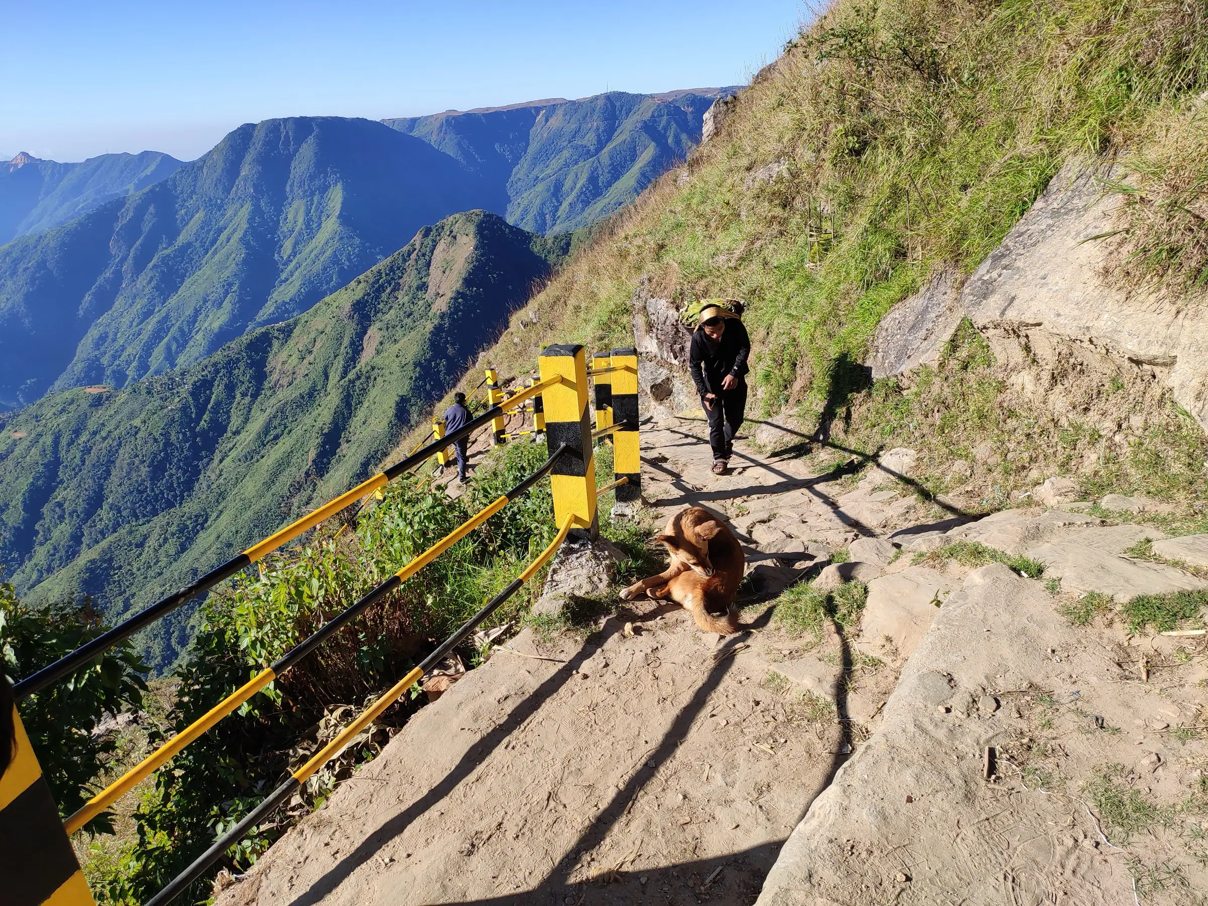 A villager carrying a load up the stone stairway with yellow-and-black railings above the Laitlum gorge, a dog resting on the steps