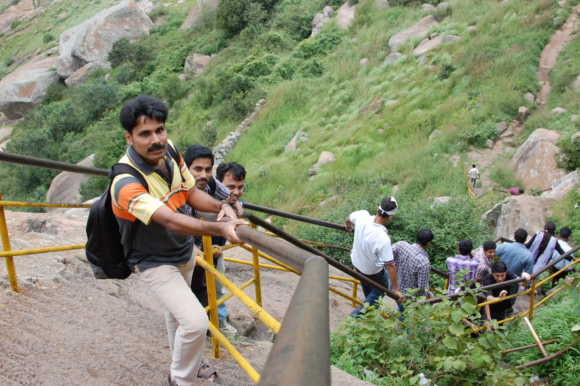 Pilgrims climbing the stepped path up Shivagange hill, holding yellow iron railings between granite boulders and grass