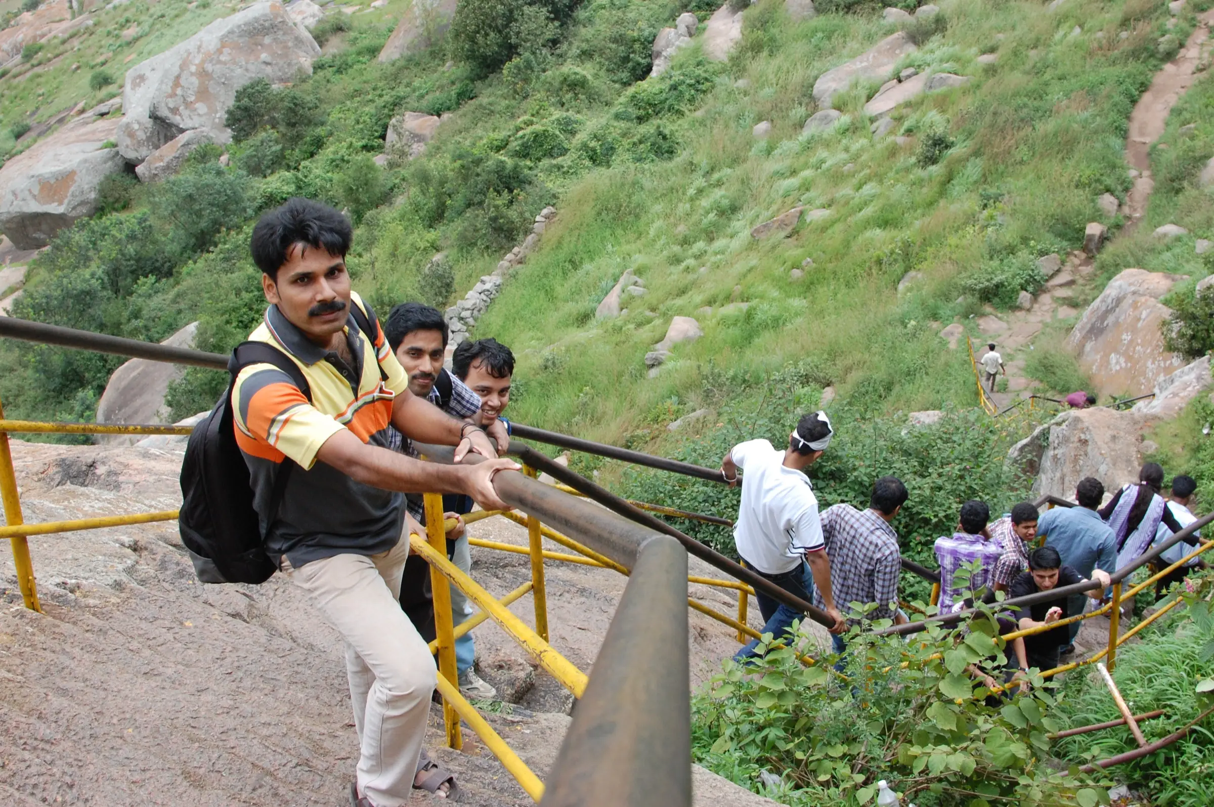 Pilgrims climbing the stepped path up Shivagange hill, holding yellow iron railings between granite boulders and grass