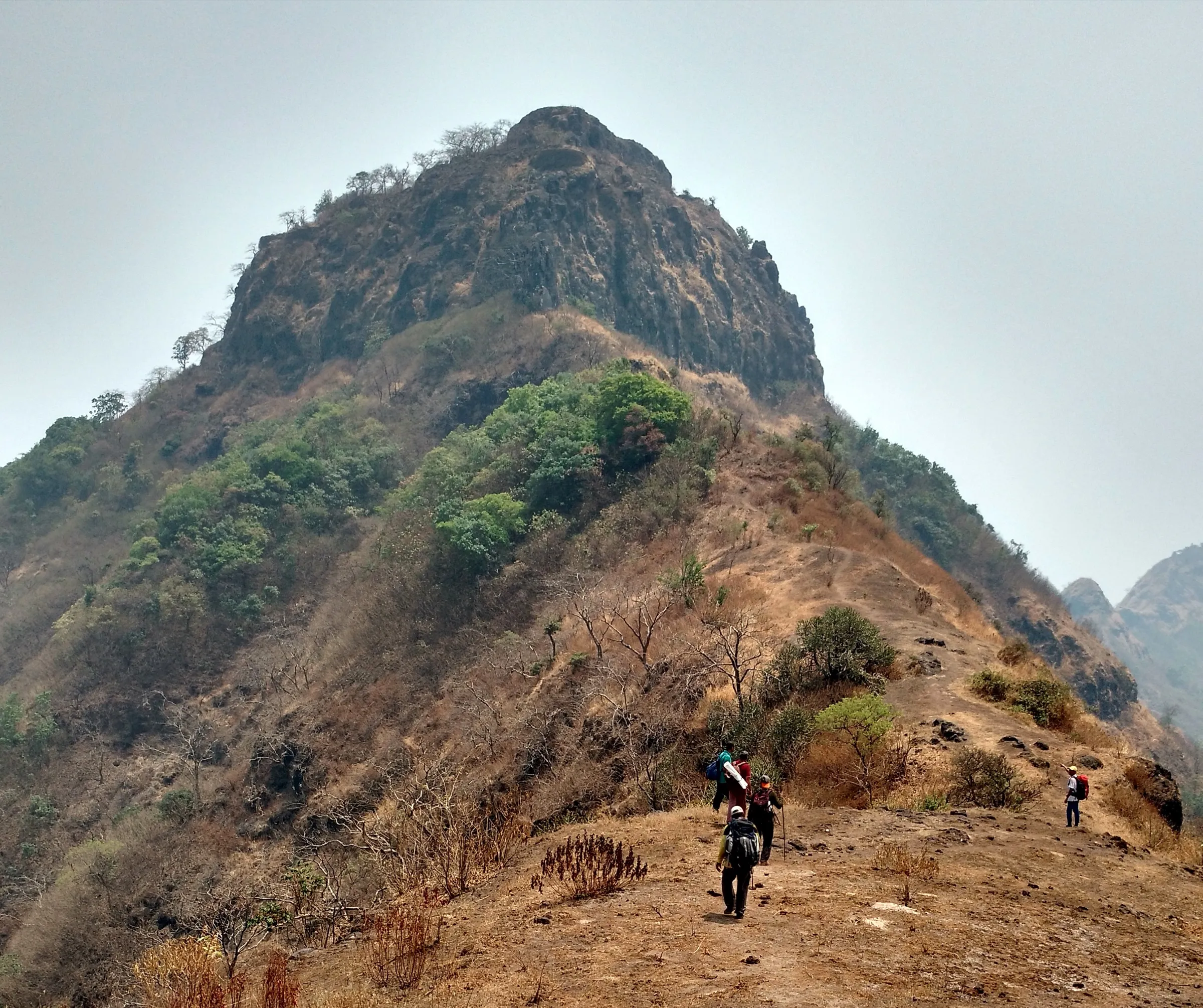 Trekkers walking a dry grassy ridge towards the rocky summit of Sumargad fort