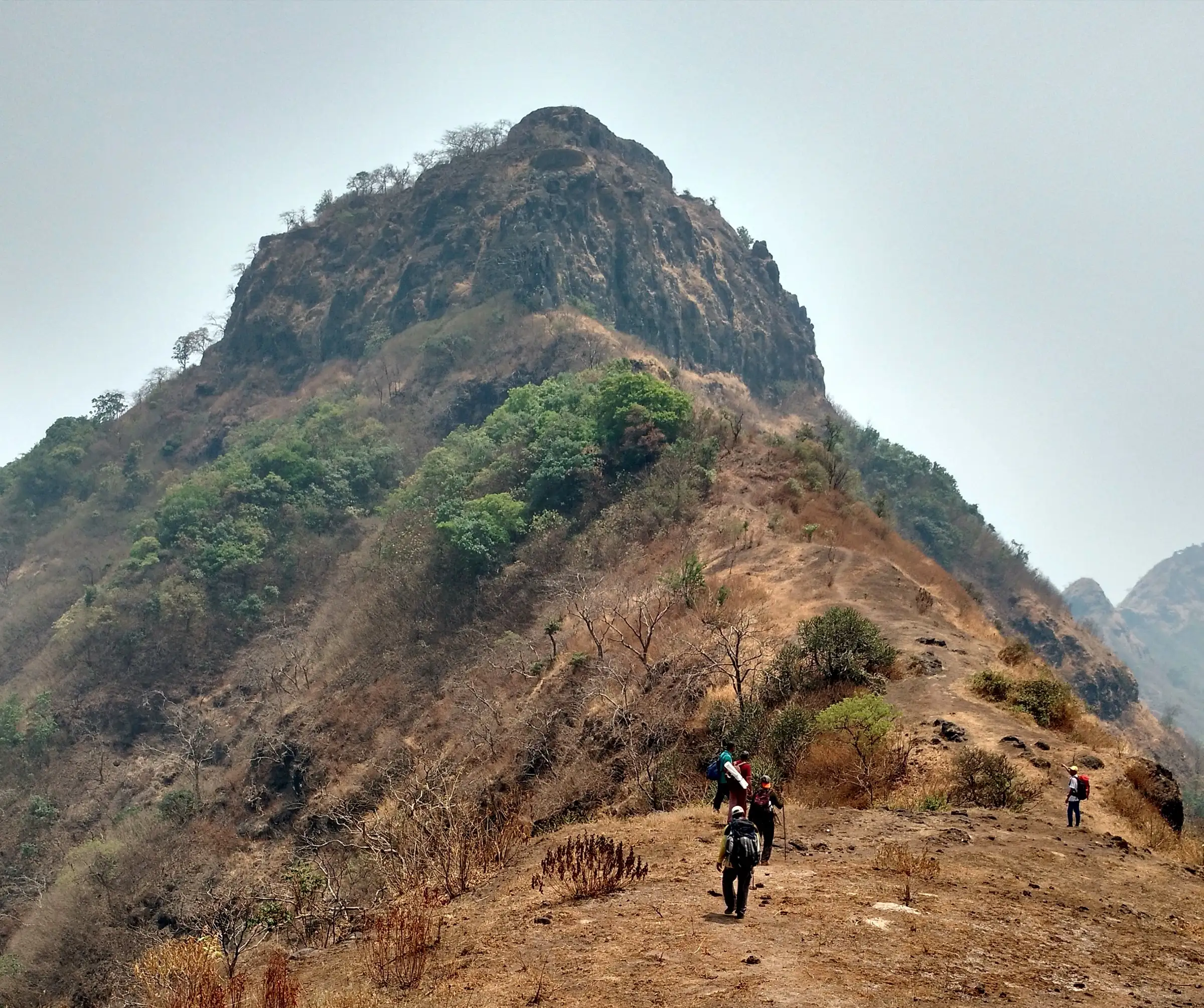 Trekkers walking a dry grassy ridge towards the rocky summit of Sumargad fort