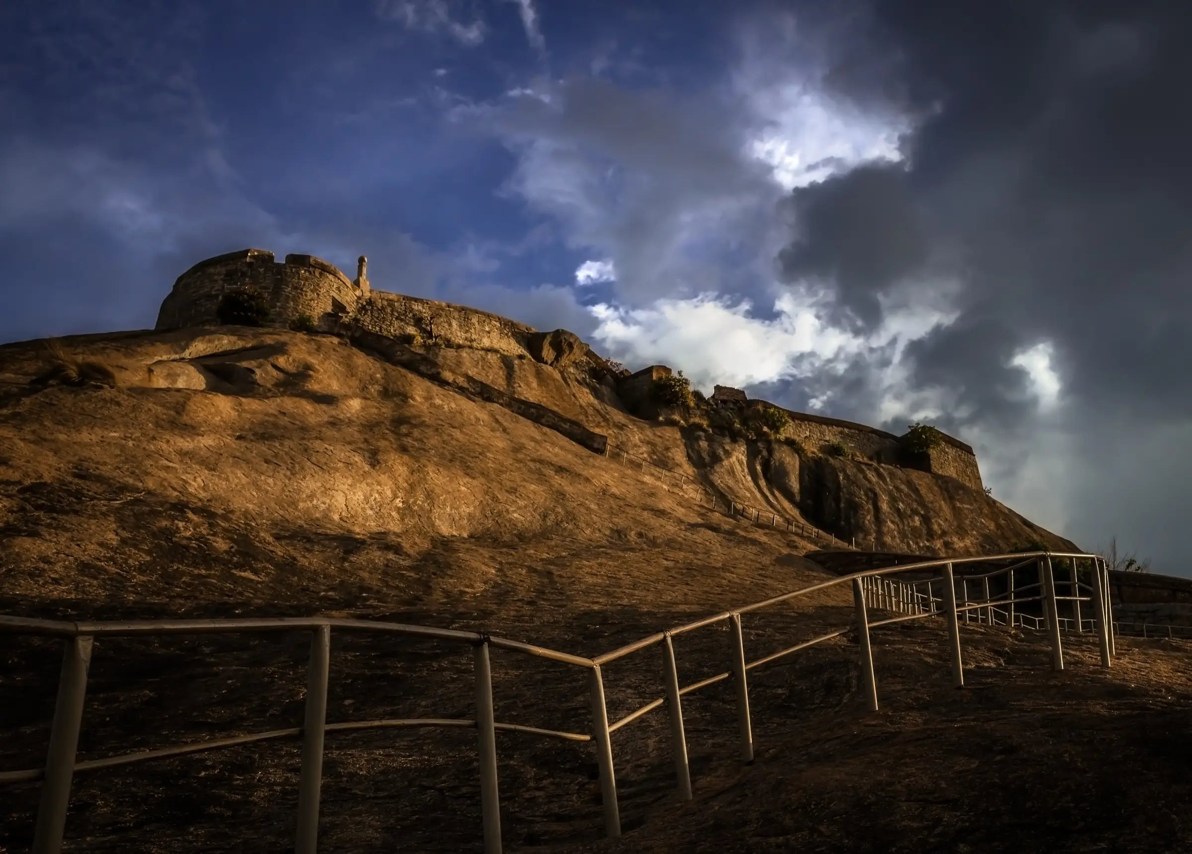 Steel railings on the bare rock below the summit fort of Madhugiri at sunset under stormy clouds