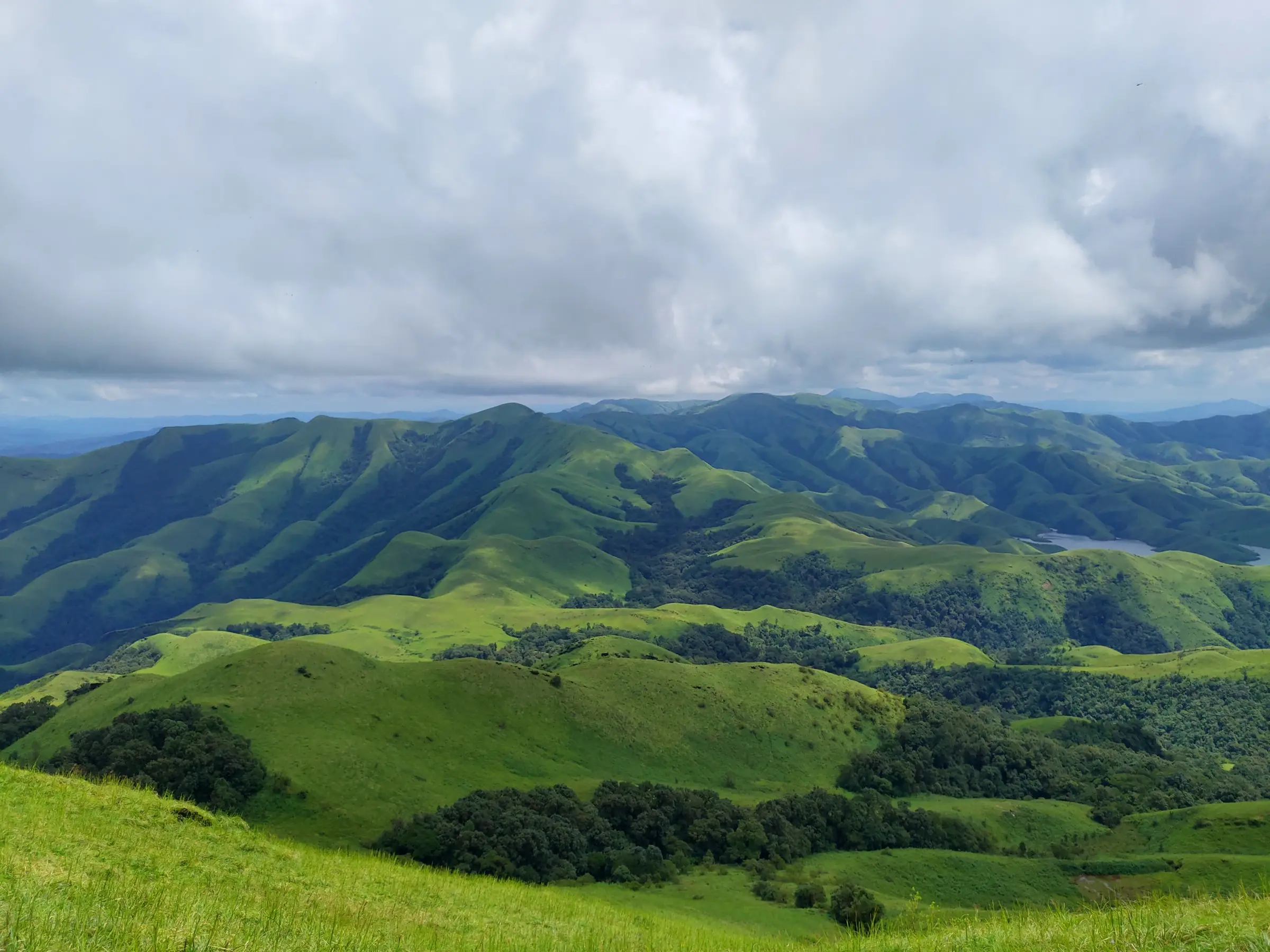 Green grassy ridges and shola forest of the Kudremukh range seen from Gangadikal under heavy cloud, with Lakya Dam backwaters at right