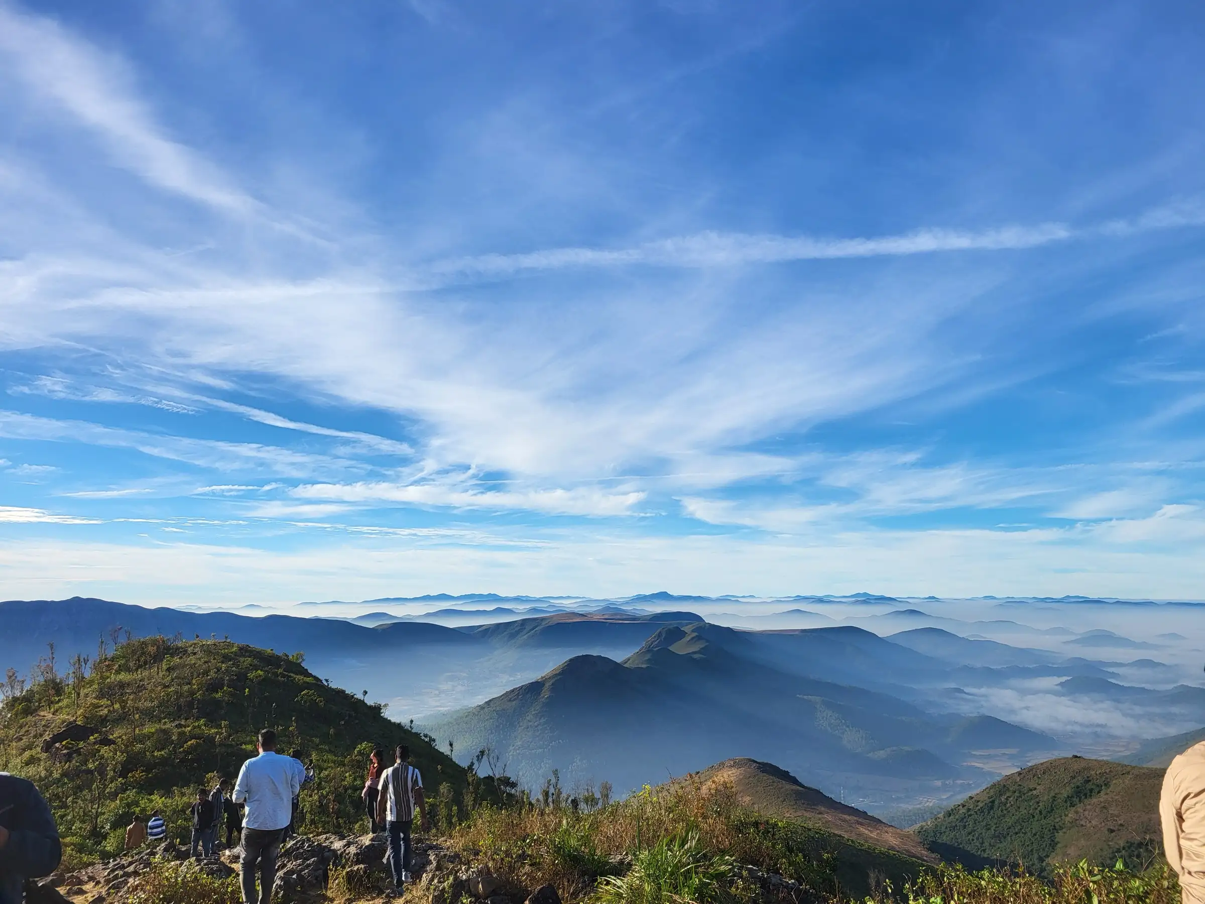 Visitors on the rocky Deomali hilltop looking out over cloud-filled valleys and layered Eastern Ghats ridges under a blue sky