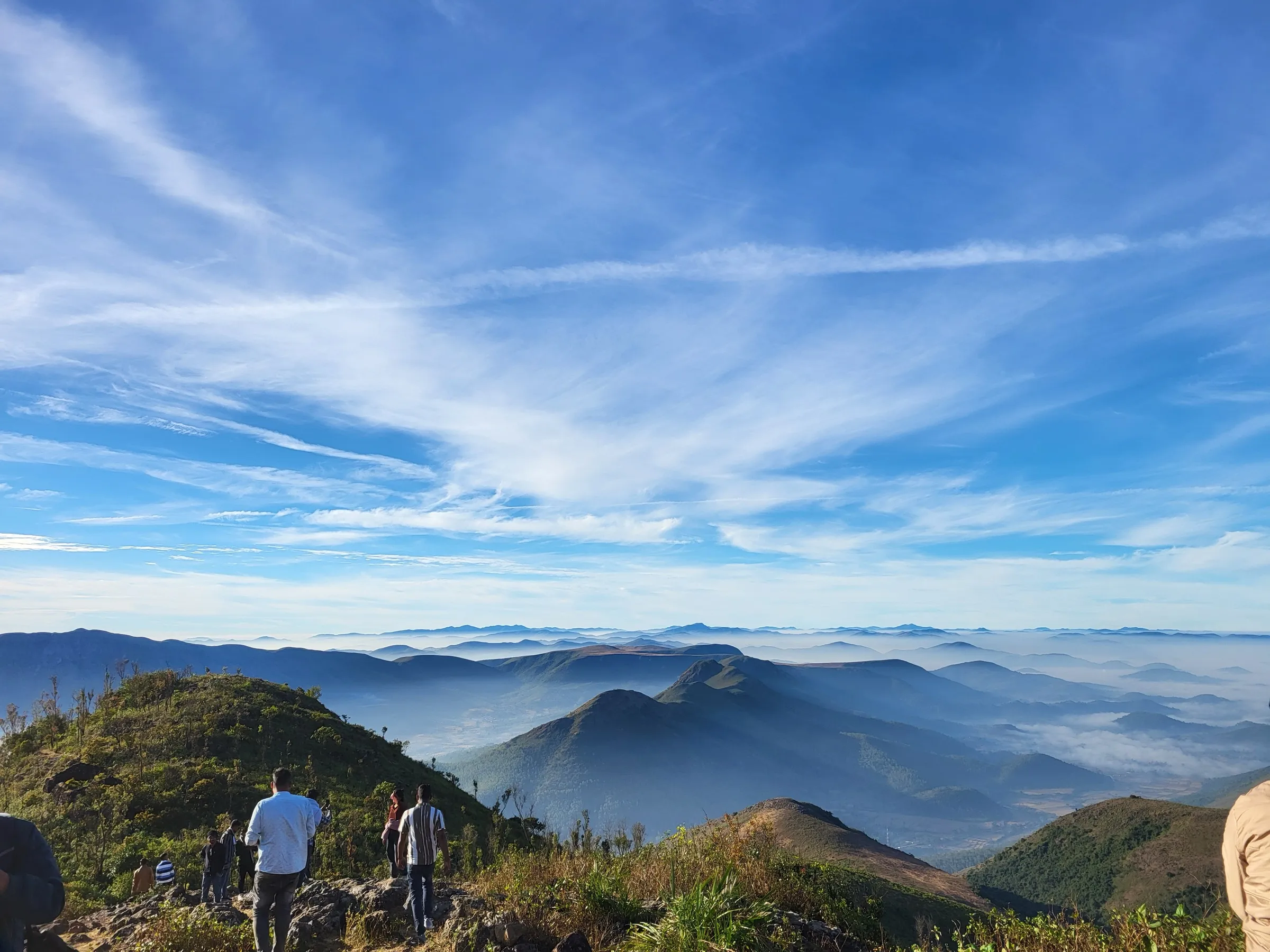 Visitors on the rocky Deomali hilltop looking out over cloud-filled valleys and layered Eastern Ghats ridges under a blue sky