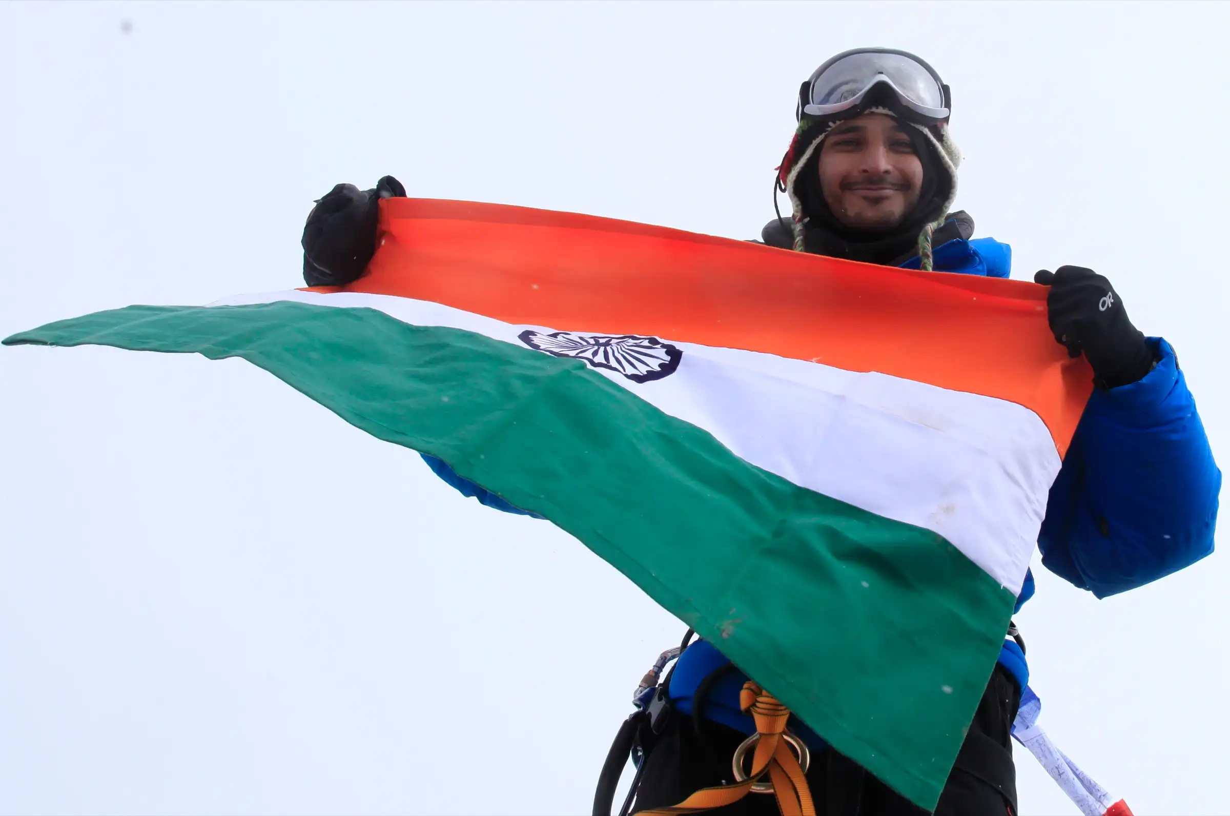 A climber in goggles and a blue down jacket holding the Indian flag on the snowy summit of Friendship Peak