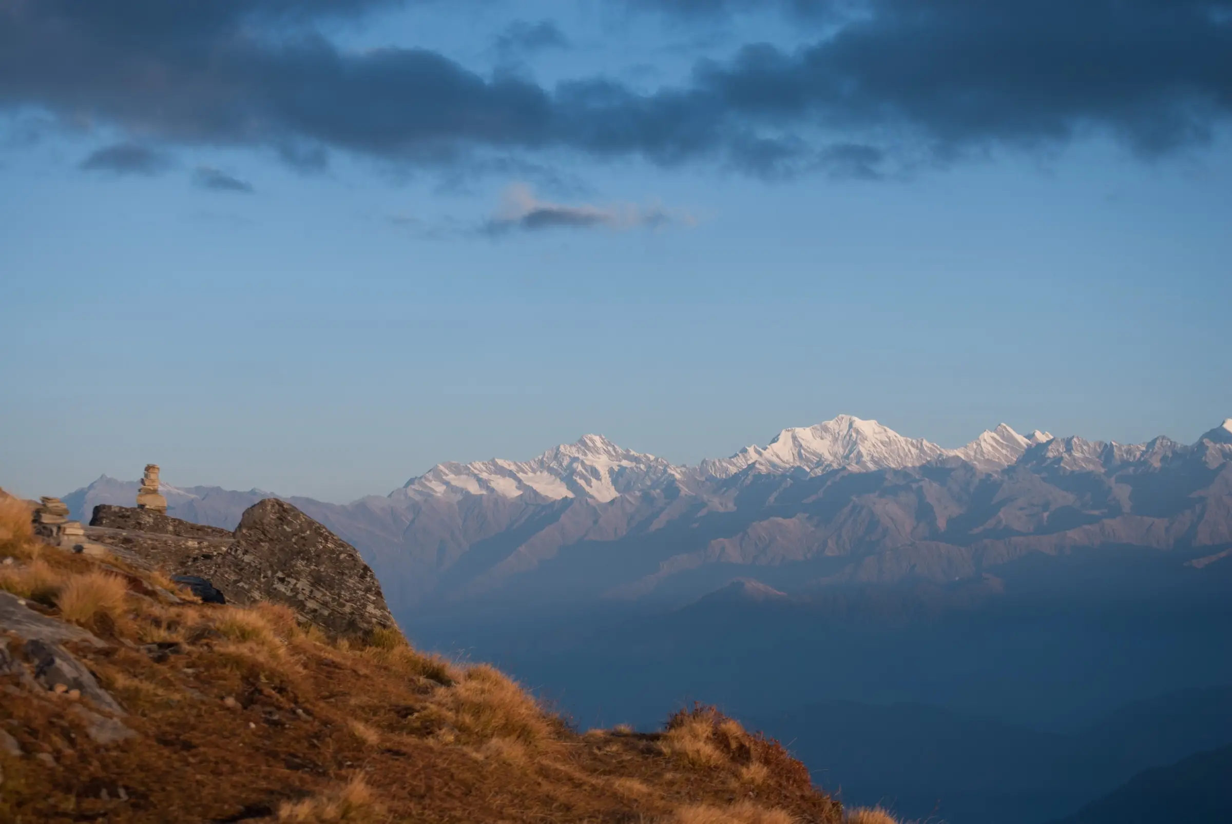 Stone cairns on the grassy Chandrashila summit with snow peaks of the Garhwal Himalaya lit by the evening sun