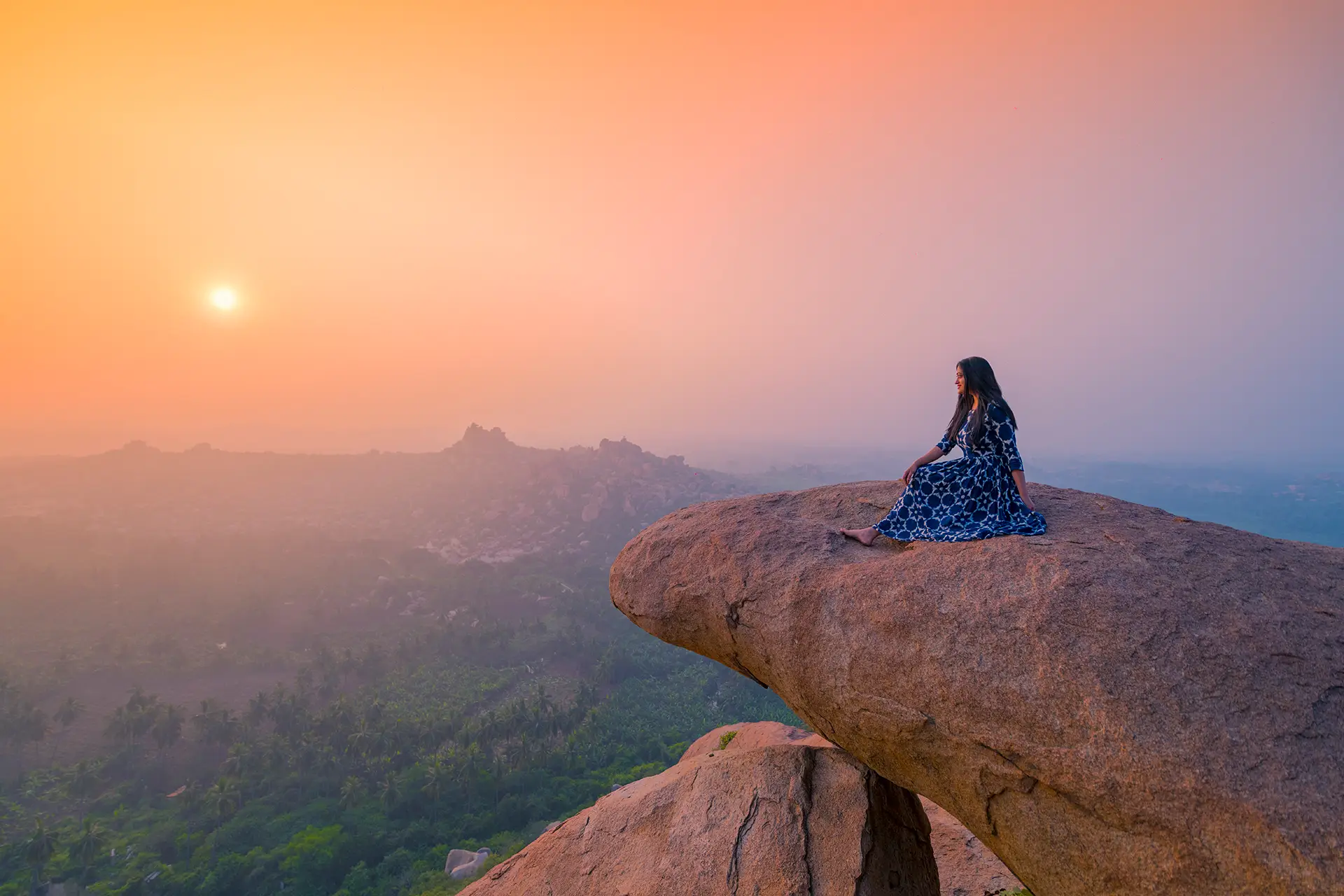 A visitor sitting on an overhanging boulder on top of Matanga Hill as the sun rises over a hazy Hampi landscape