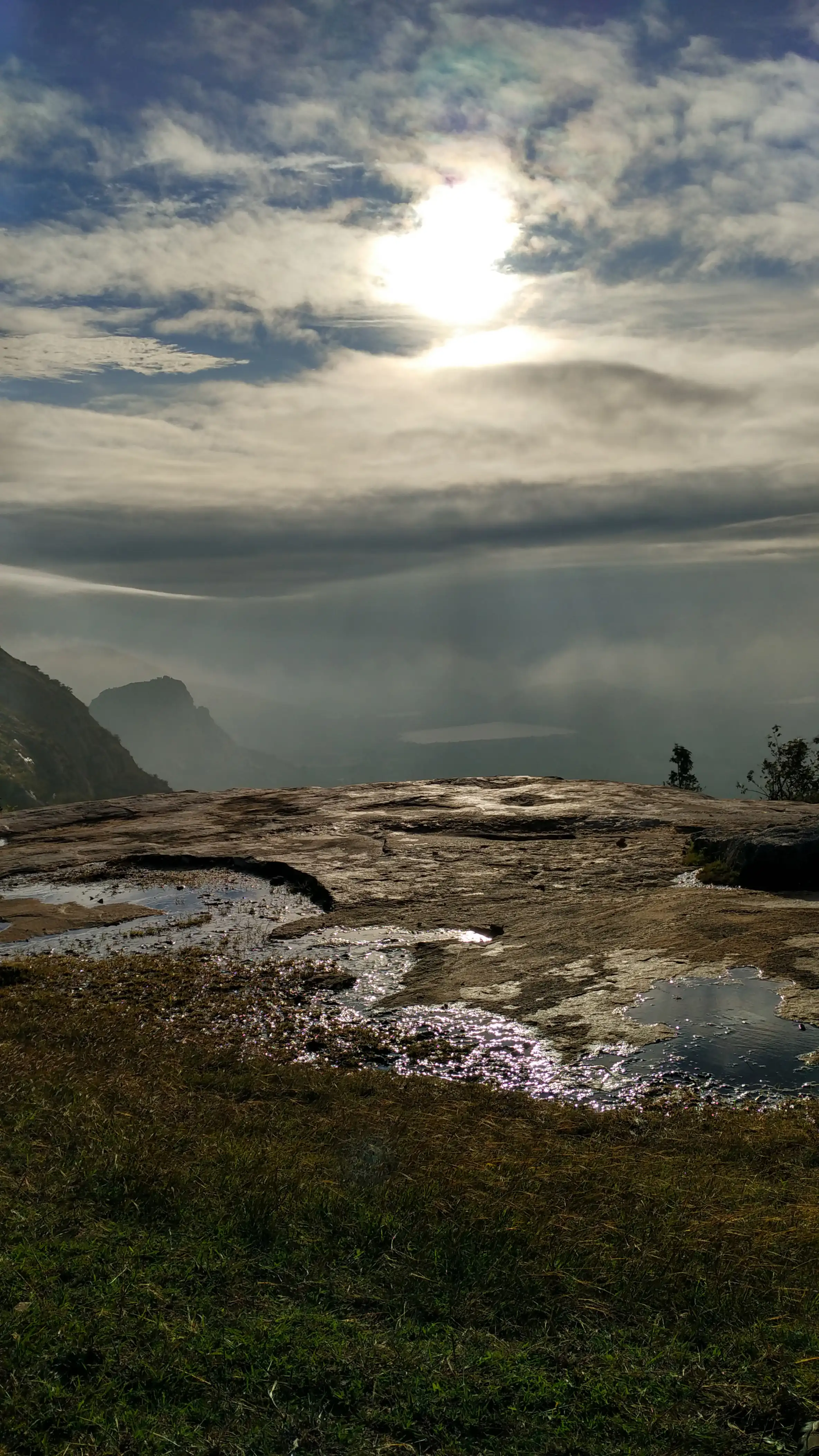 Morning sun breaking through cloud over a granite hilltop at Kaiwara, with rain-filled rock pools and distant hills in haze