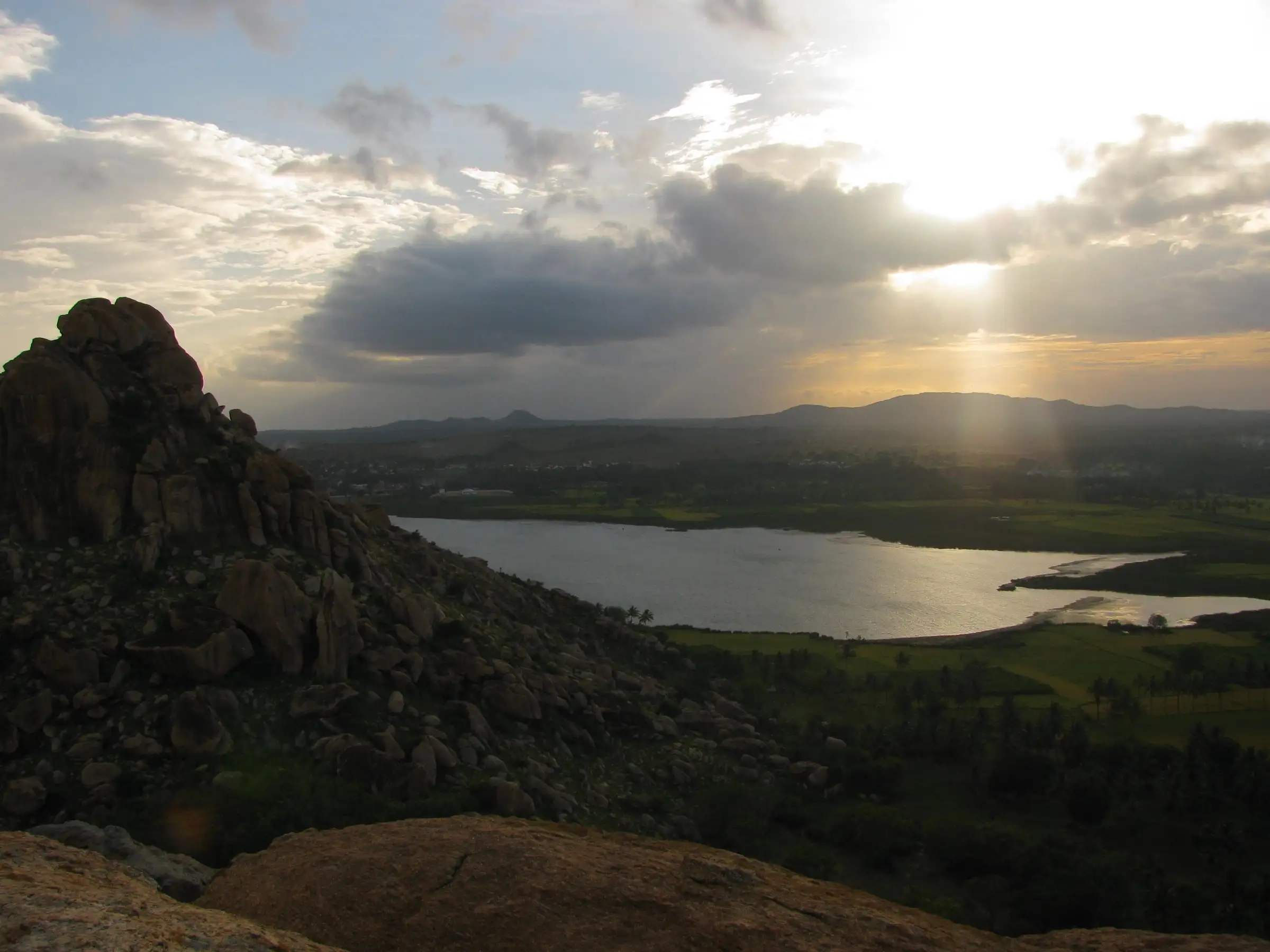 Sun breaking through clouds over a lake and farmland seen from the granite boulders of Kunti Betta