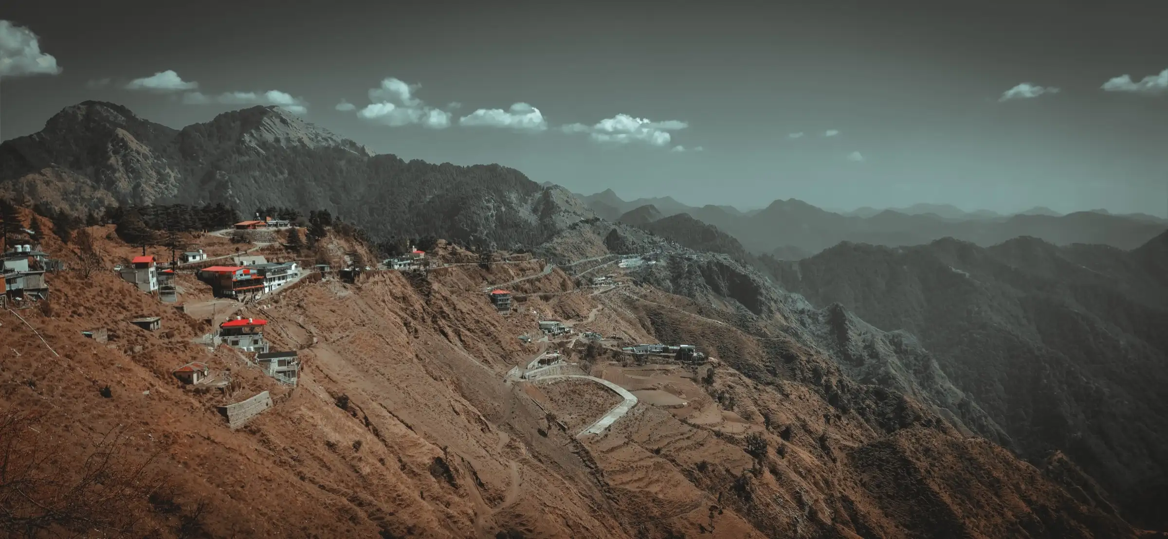 Houses and a winding road on a dry terraced hillside at Suwakholi, below forested ridges near Mussoorie