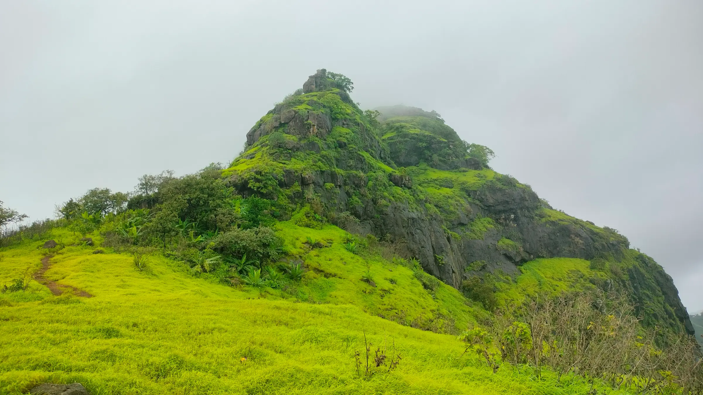 The rocky, forested hill of Takmak Fort rising above a bright green grassy slope under monsoon cloud