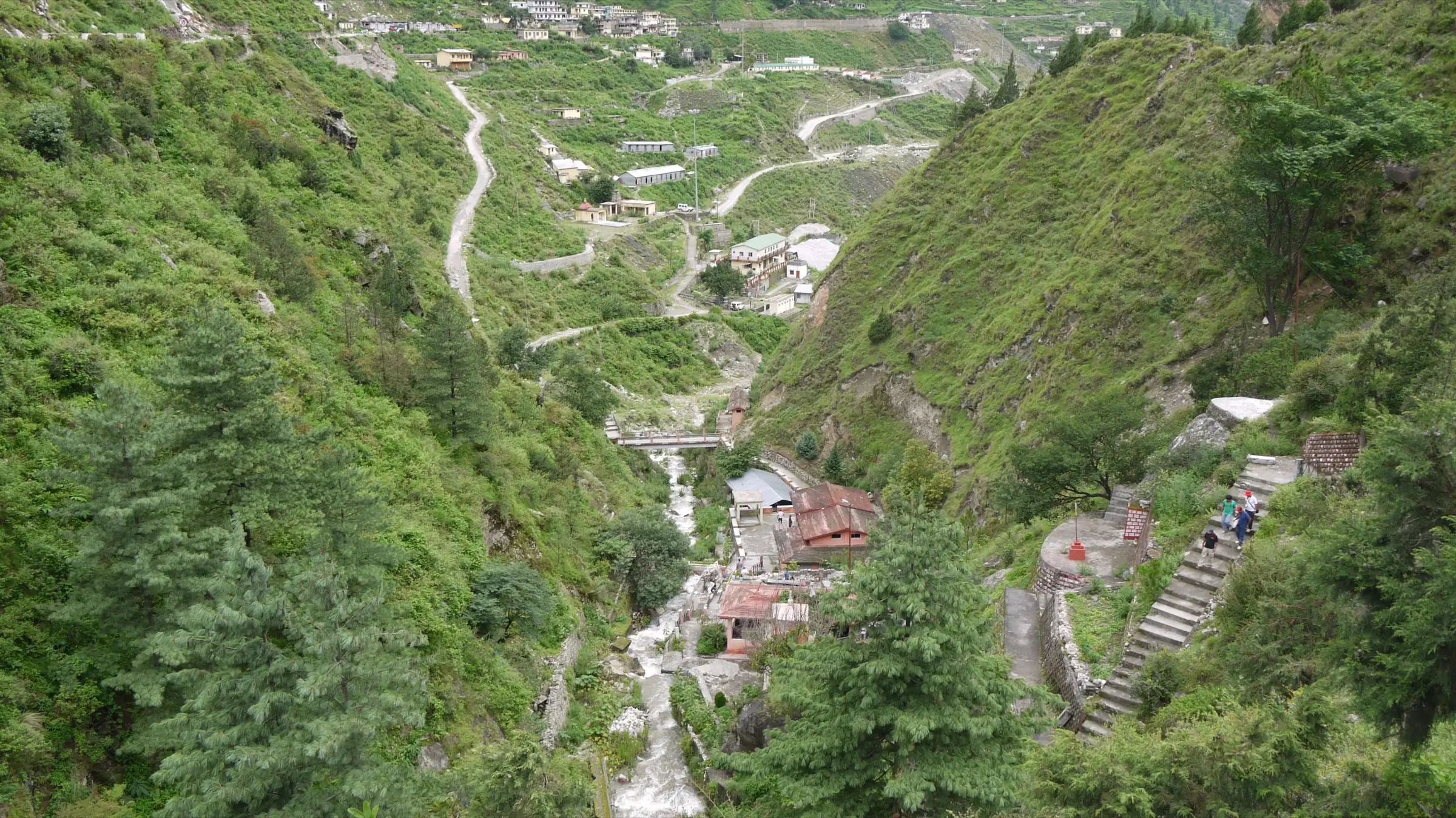 A road zigzagging up green slopes above a stream and buildings at Tapovan near Joshimath, close to the Saldhar trailhead