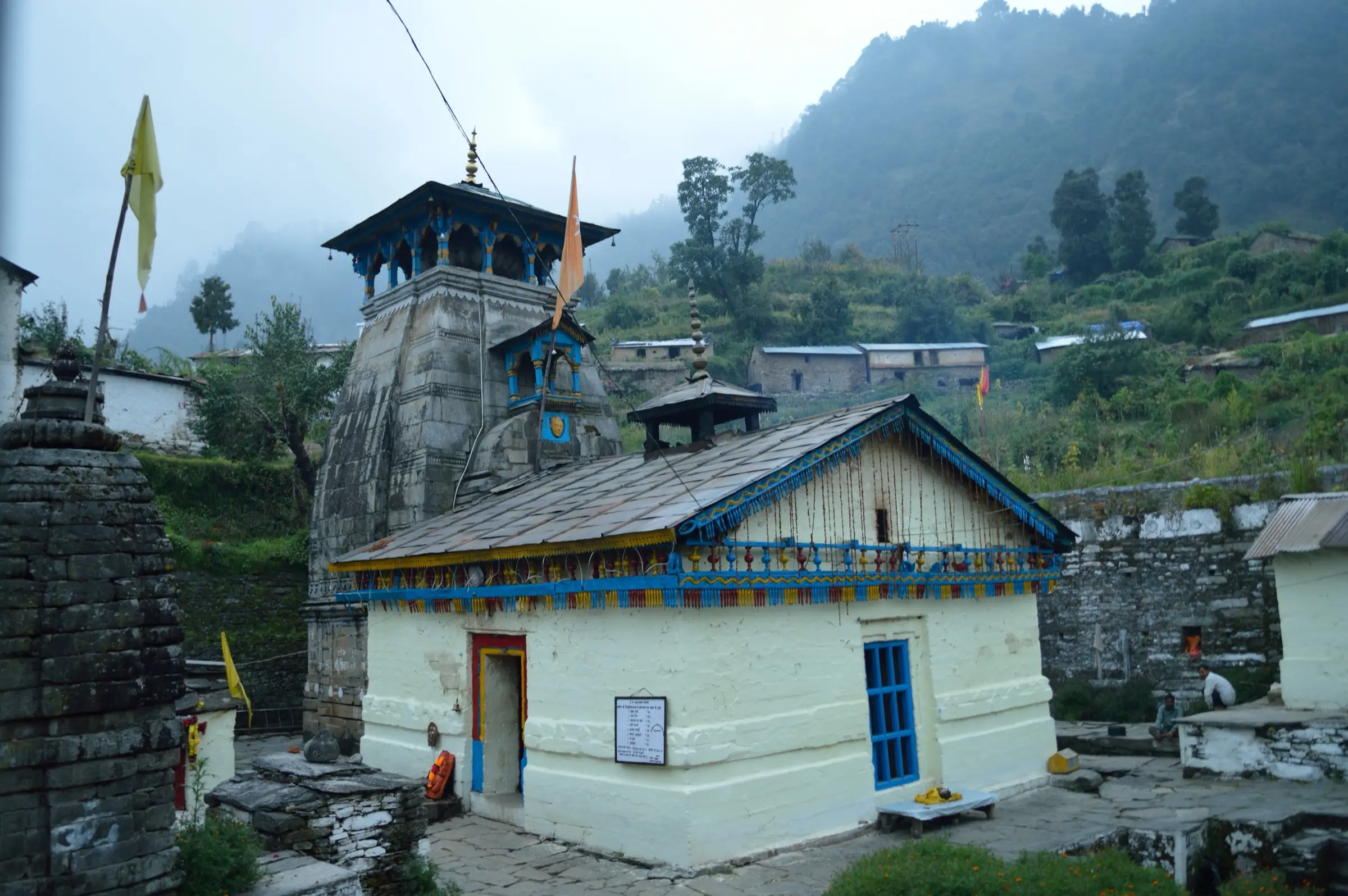 Triyuginarayan temple on a misty October day, with saffron flags beside the stone shikhar and forested hills behind the village
