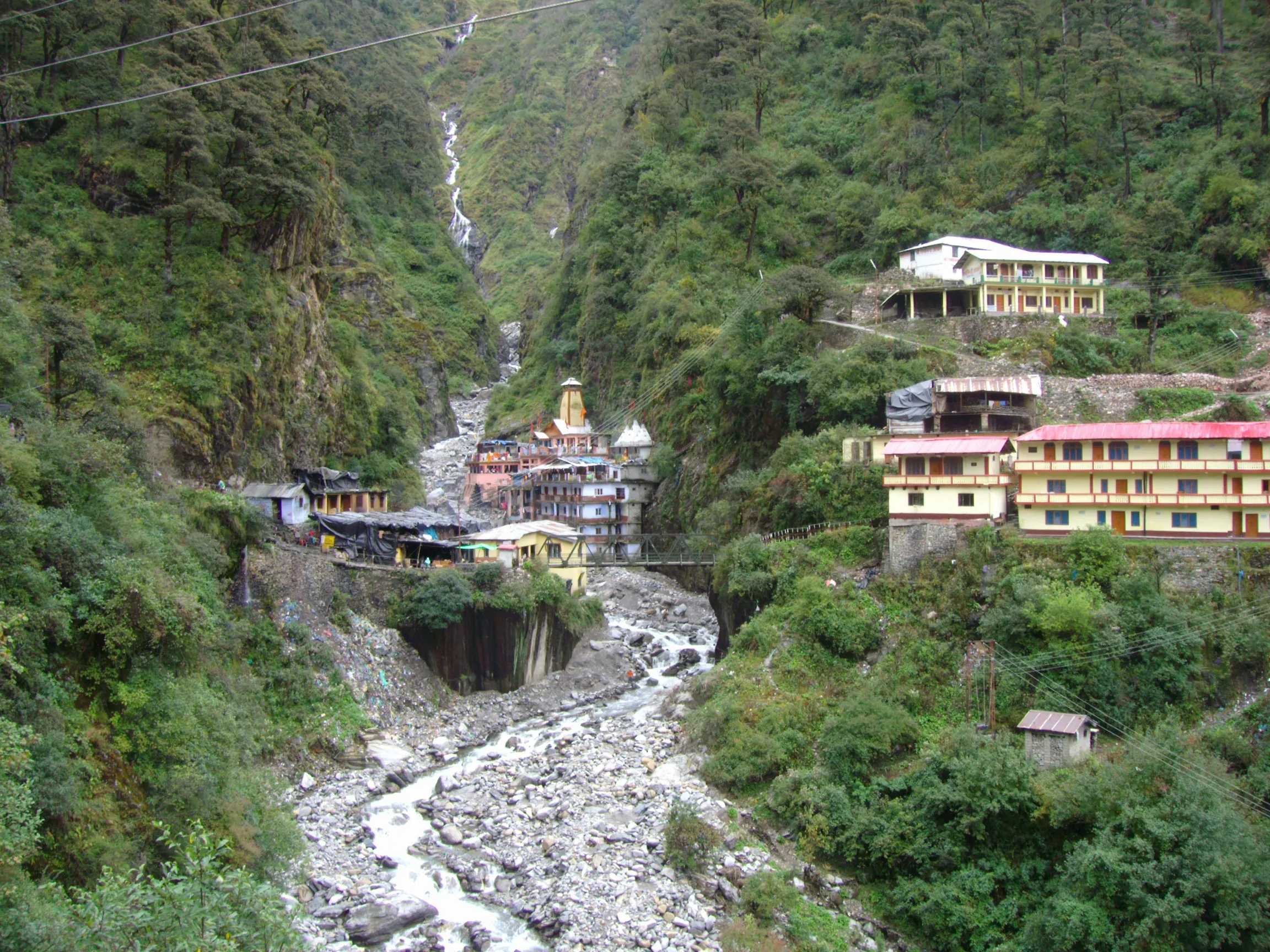 Yamunotri temple and ashram buildings in a green, forested gorge above the rocky bed of the Yamuna, with a waterfall behind