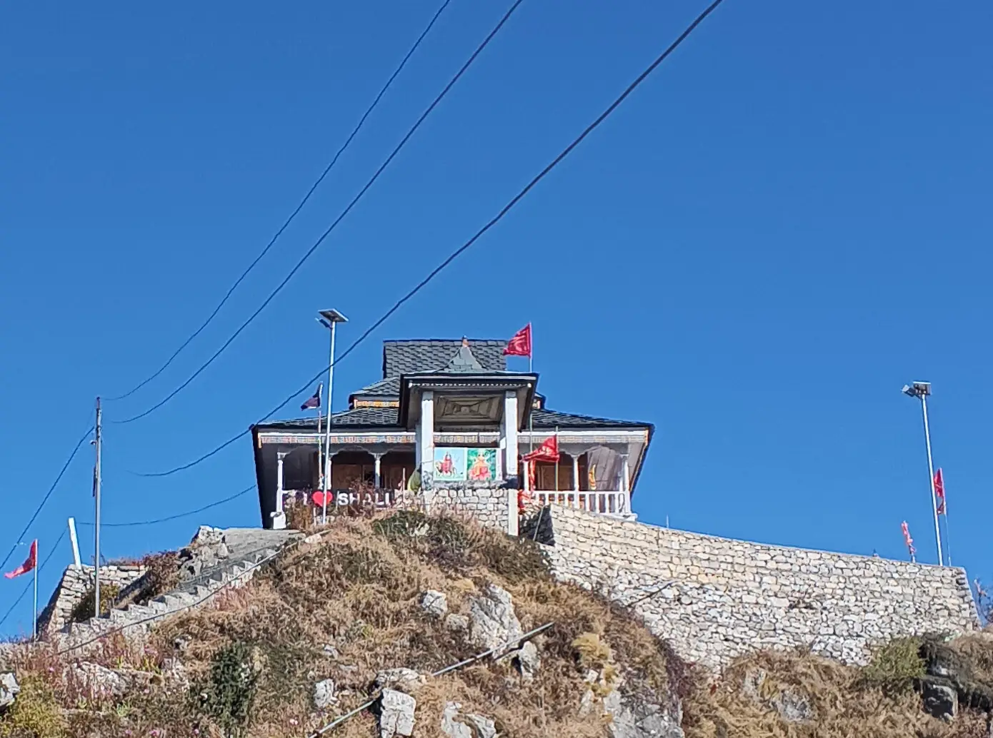 Close view of the wooden Shali Mata Temple with a slate roof and red flags on a stone retaining wall atop Shali Tibba