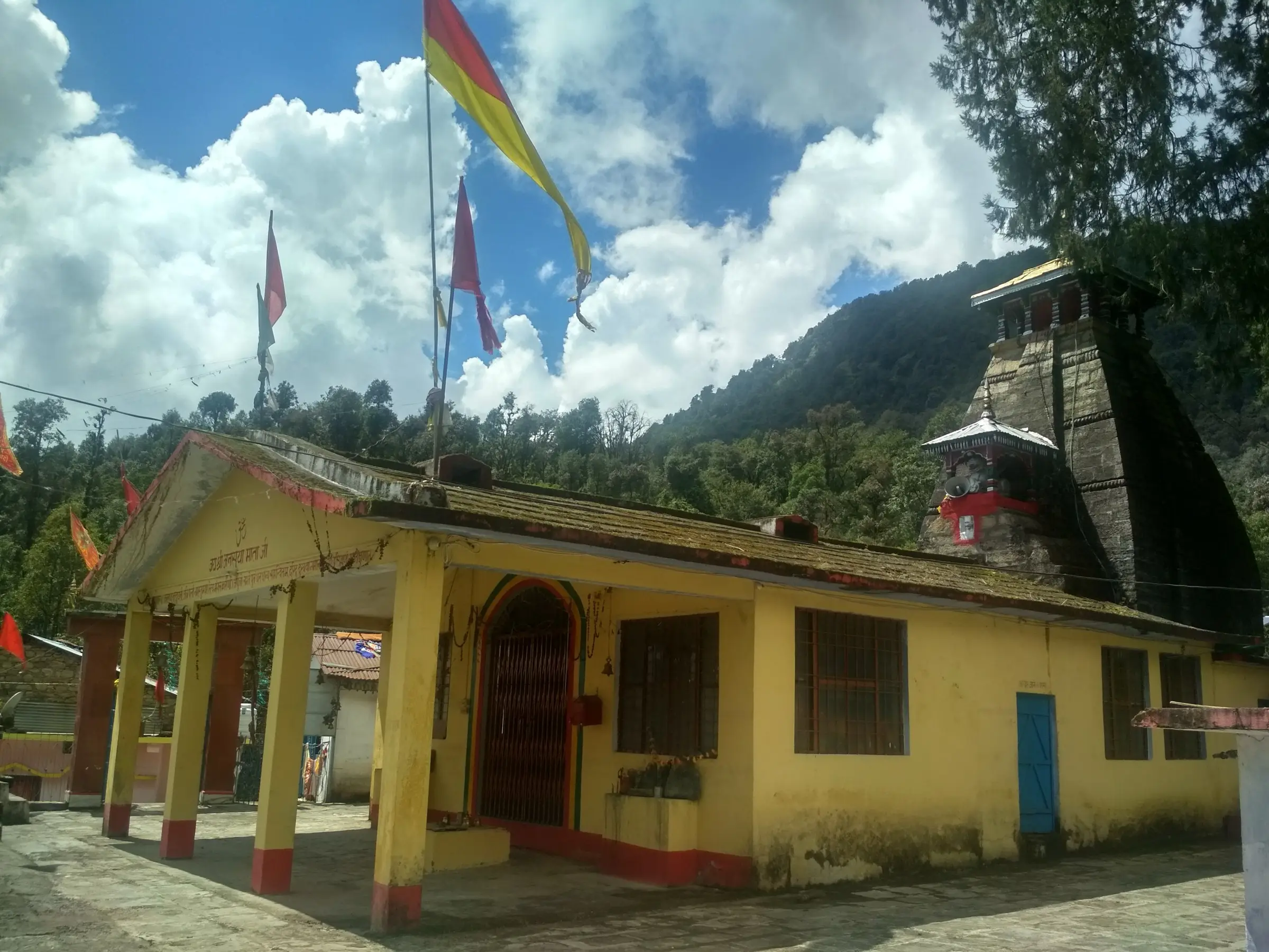 Yellow mandap and porch of the Anusuya Mata temple with saffron flags and the dark stone shikhara behind