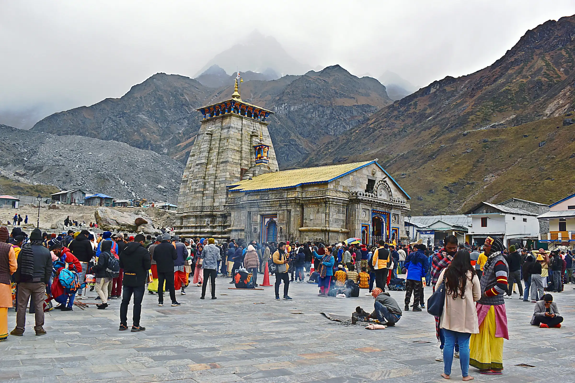 Devotees on the stone-paved courtyard in front of Kedarnath temple, with rocky mountains and cloud-covered snow peaks behind