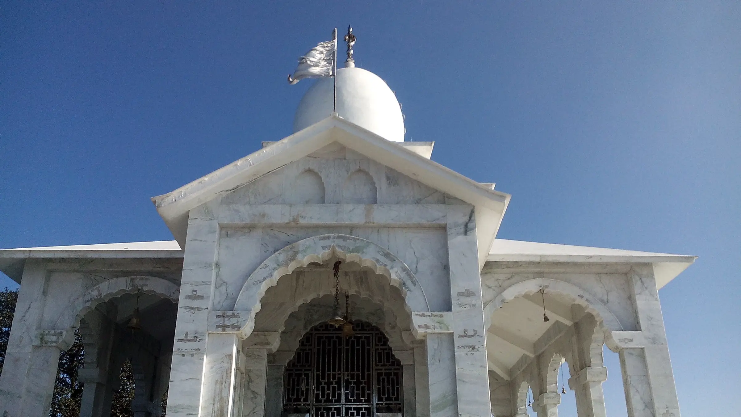 Front of the marble Bhadraj Temple with carved arches, a saffron-white flag and the domed shikhara
