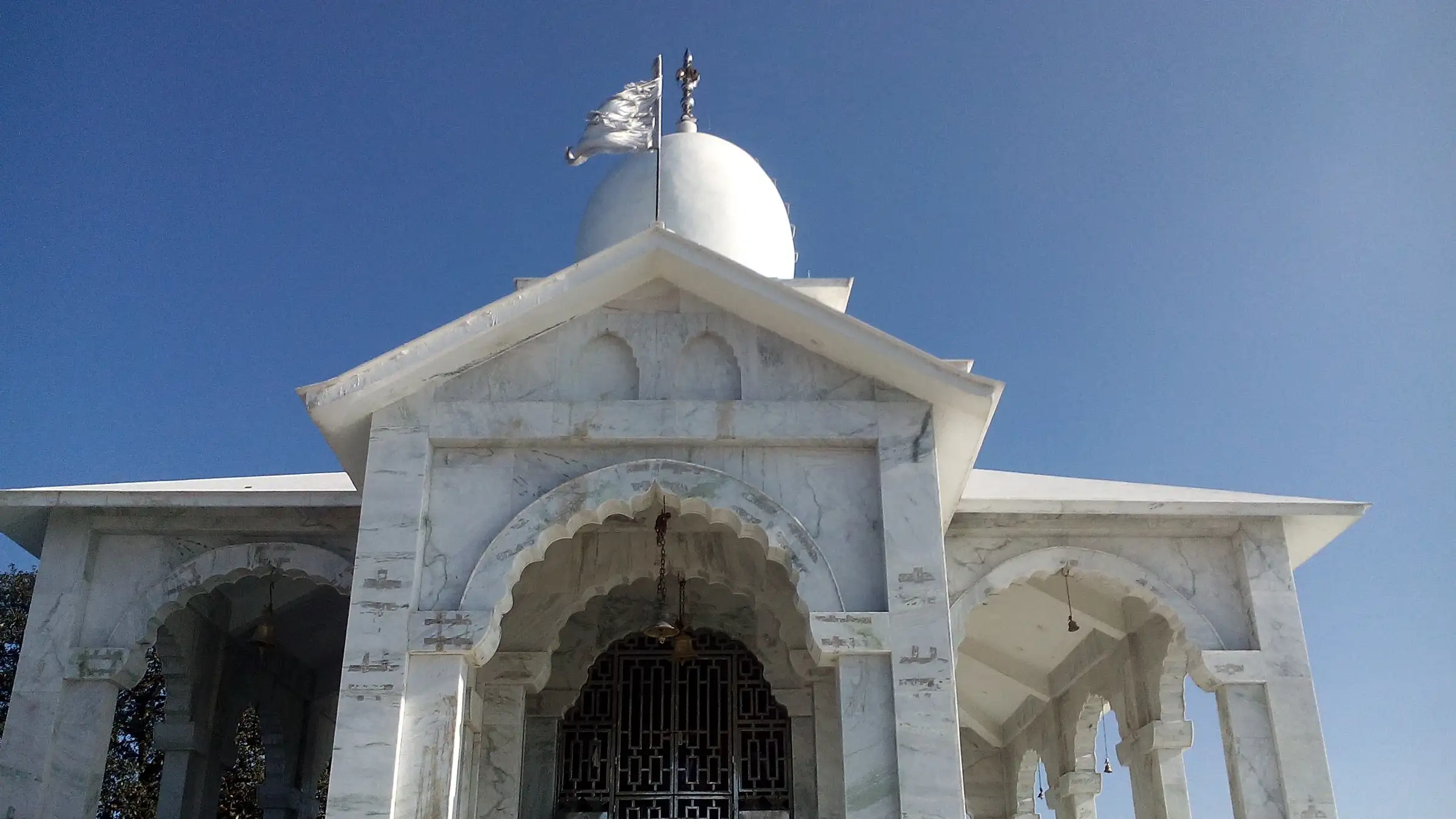 Front of the marble Bhadraj Temple with carved arches, a saffron-white flag and the domed shikhara