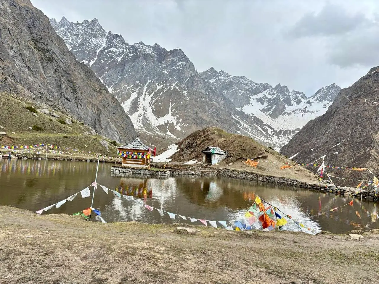 Yulla Kanda lake with the Krishna temple on its stone platform, prayer flags and a small stone hut, under jagged snowy peaks