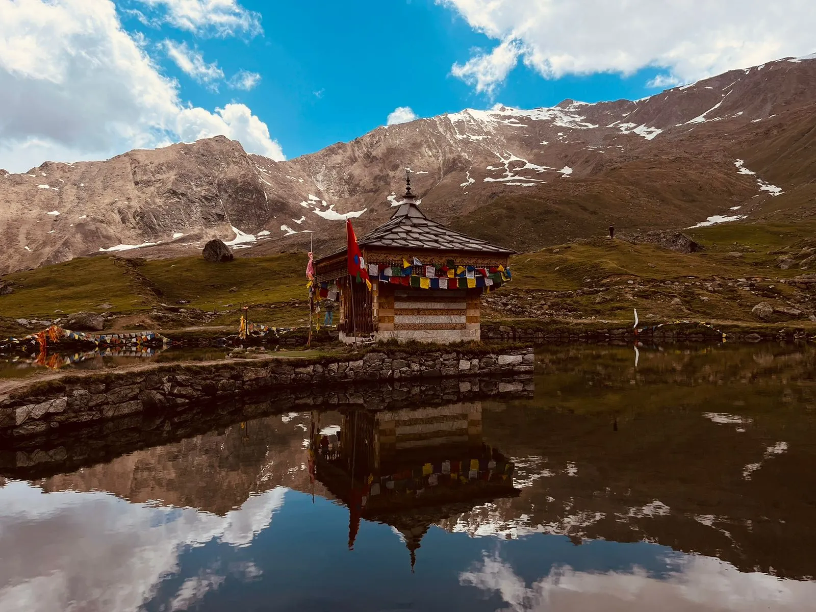 The wooden Shri Krishna temple with prayer flags reflected in the still Yulla Kanda lake, below grassy slopes and snow-streaked mountains