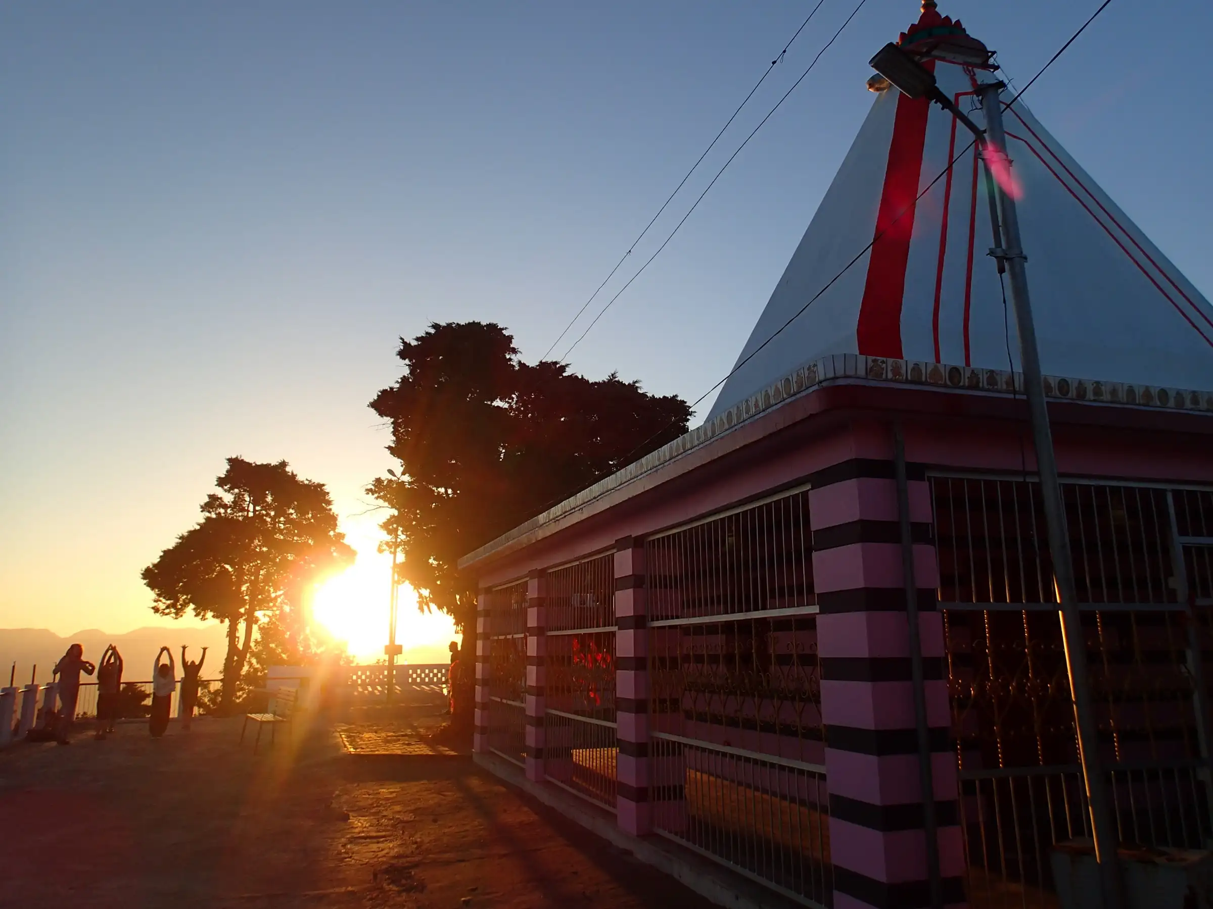 Sunrise behind trees beside the white-and-red shikhara of Kunjapuri Devi temple, with visitors on the terrace and hills on the horizon