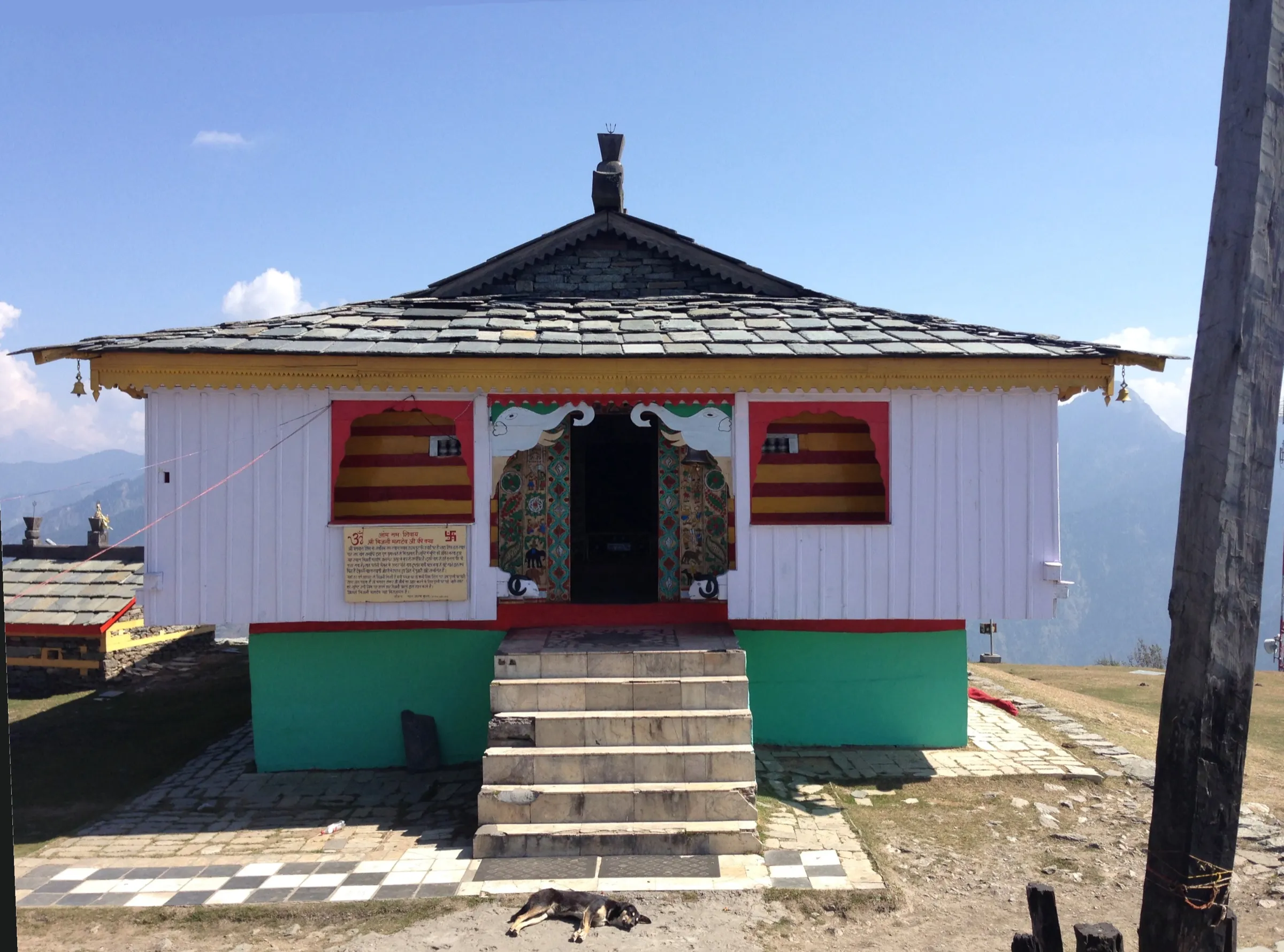 White wooden Bijli Mahadev temple with a slate roof and painted doors on the Mathan hilltop under a blue sky