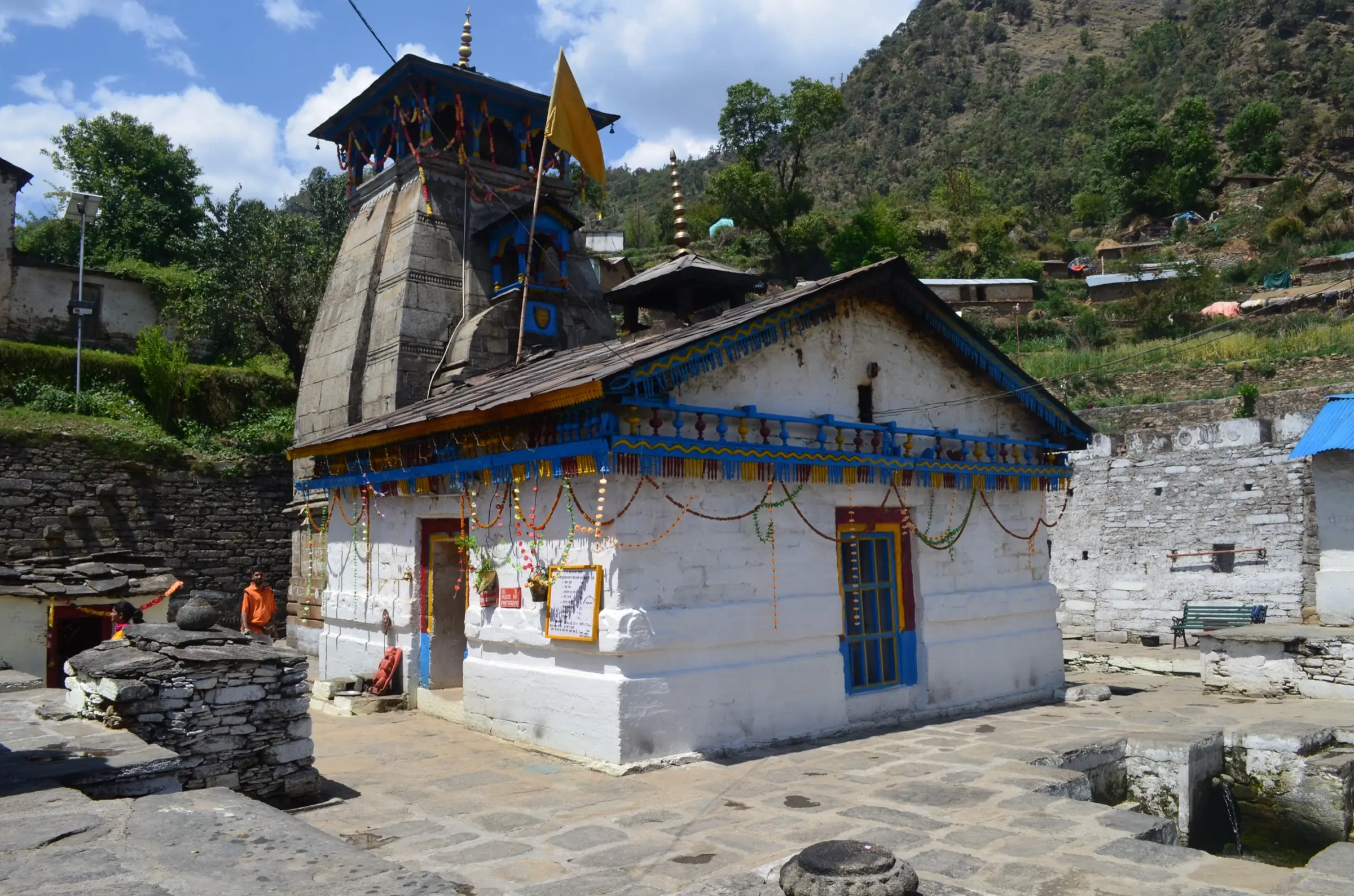 Triyuginarayan temple with its stone Kedar-style shikhar and a whitewashed mandapa decorated with blue trim and marigold garlands, under a terraced green hillside