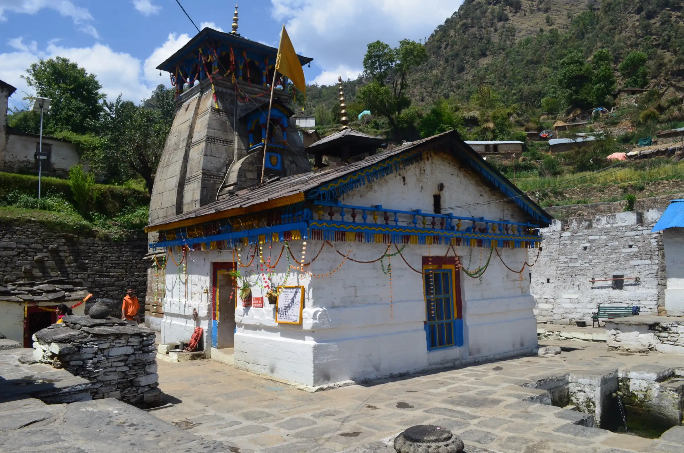 Triyuginarayan temple with its stone Kedar-style shikhar and a whitewashed mandapa decorated with blue trim and marigold garlands, under a terraced green hillside