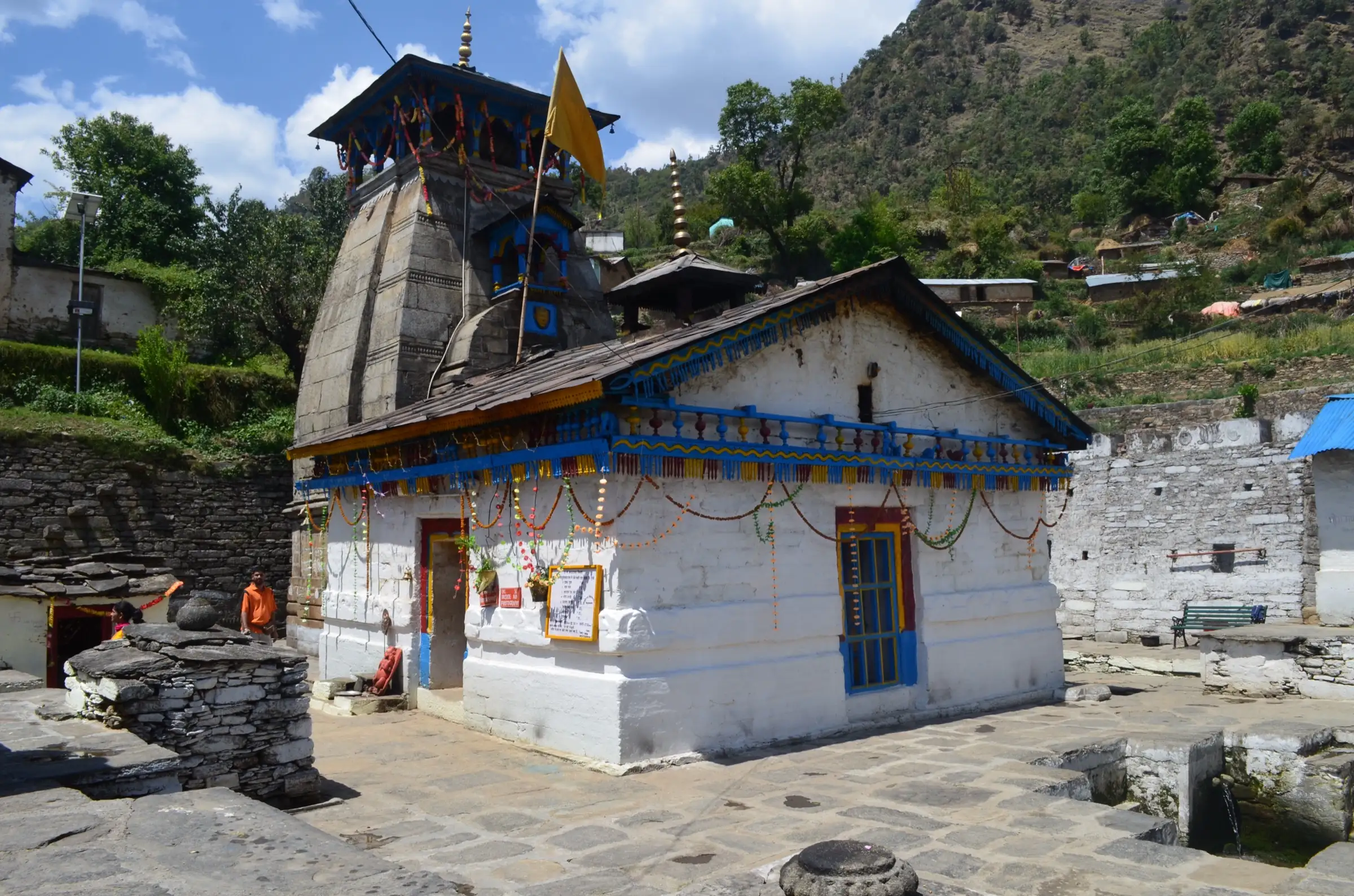 Triyuginarayan temple with its stone Kedar-style shikhar and a whitewashed mandapa decorated with blue trim and marigold garlands, under a terraced green hillside