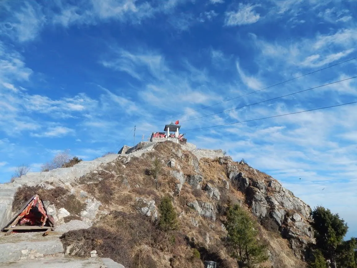 Stone steps climbing a rocky, grassy hilltop to the small Shali Mata temple with a red flag on the summit of Shali Tibba under a blue sky