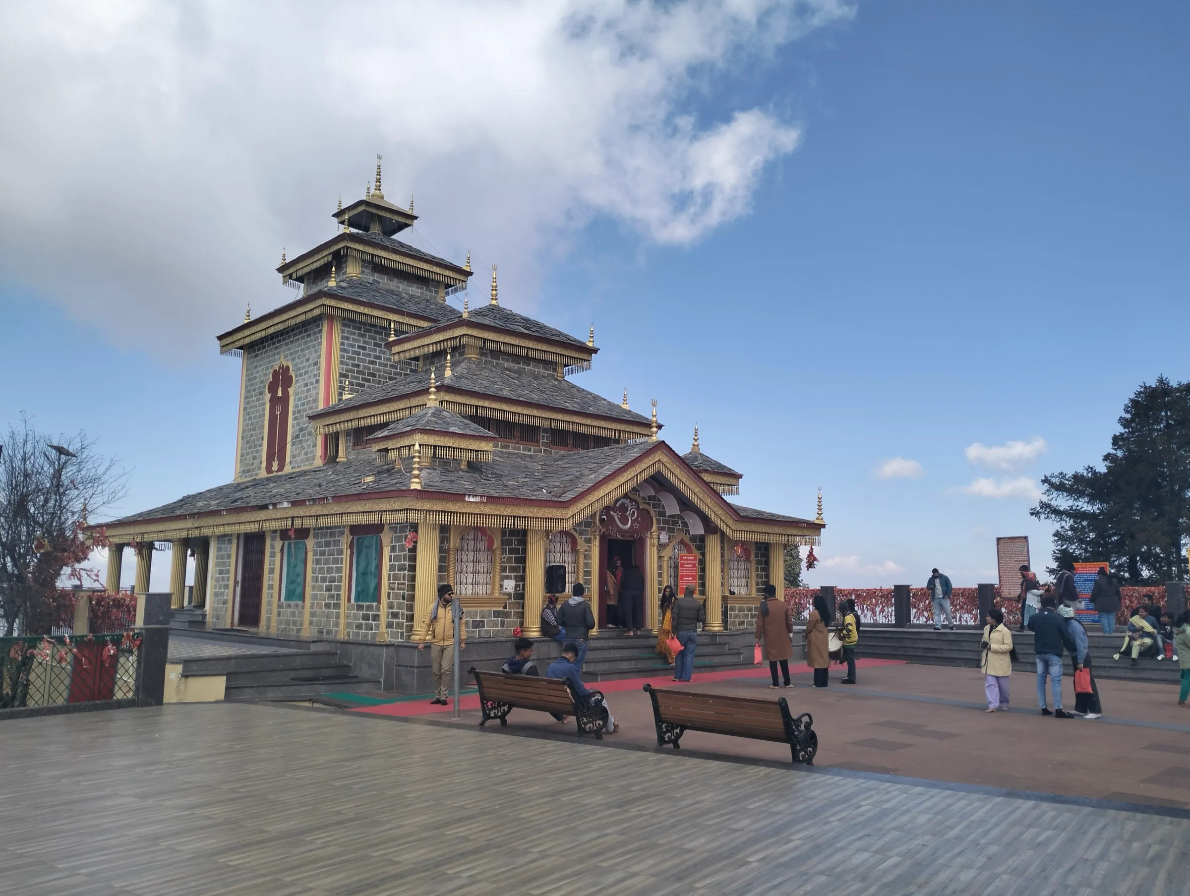 Surkanda Devi temple with its tiered pagoda-style roof on the paved hilltop courtyard, devotees and benches under a blue sky