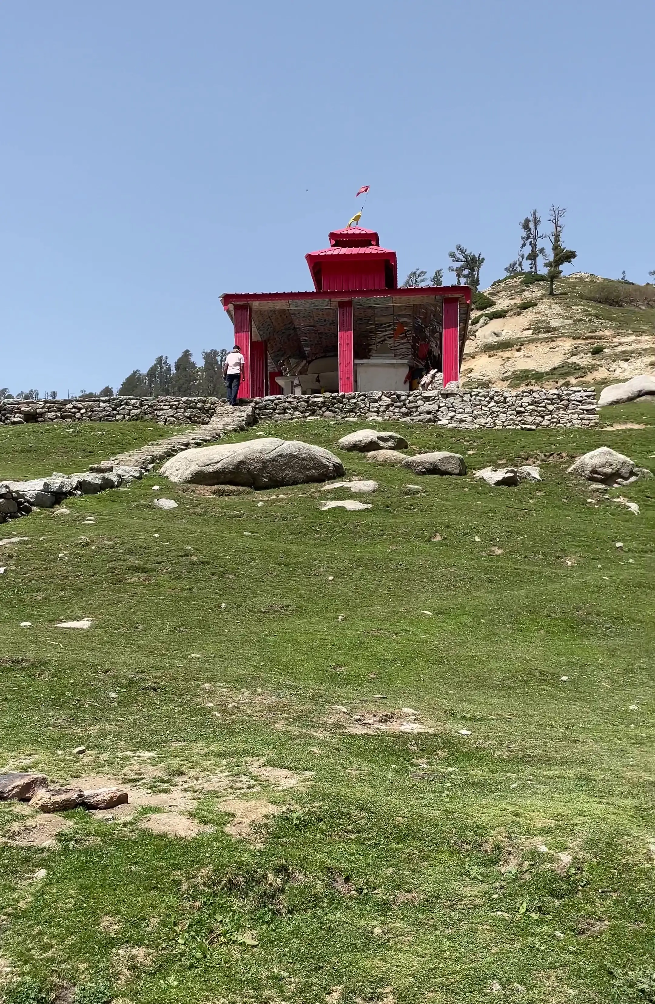 Red Shiv-Parvati temple on a grassy hilltop at Kareri Lake under a clear sky