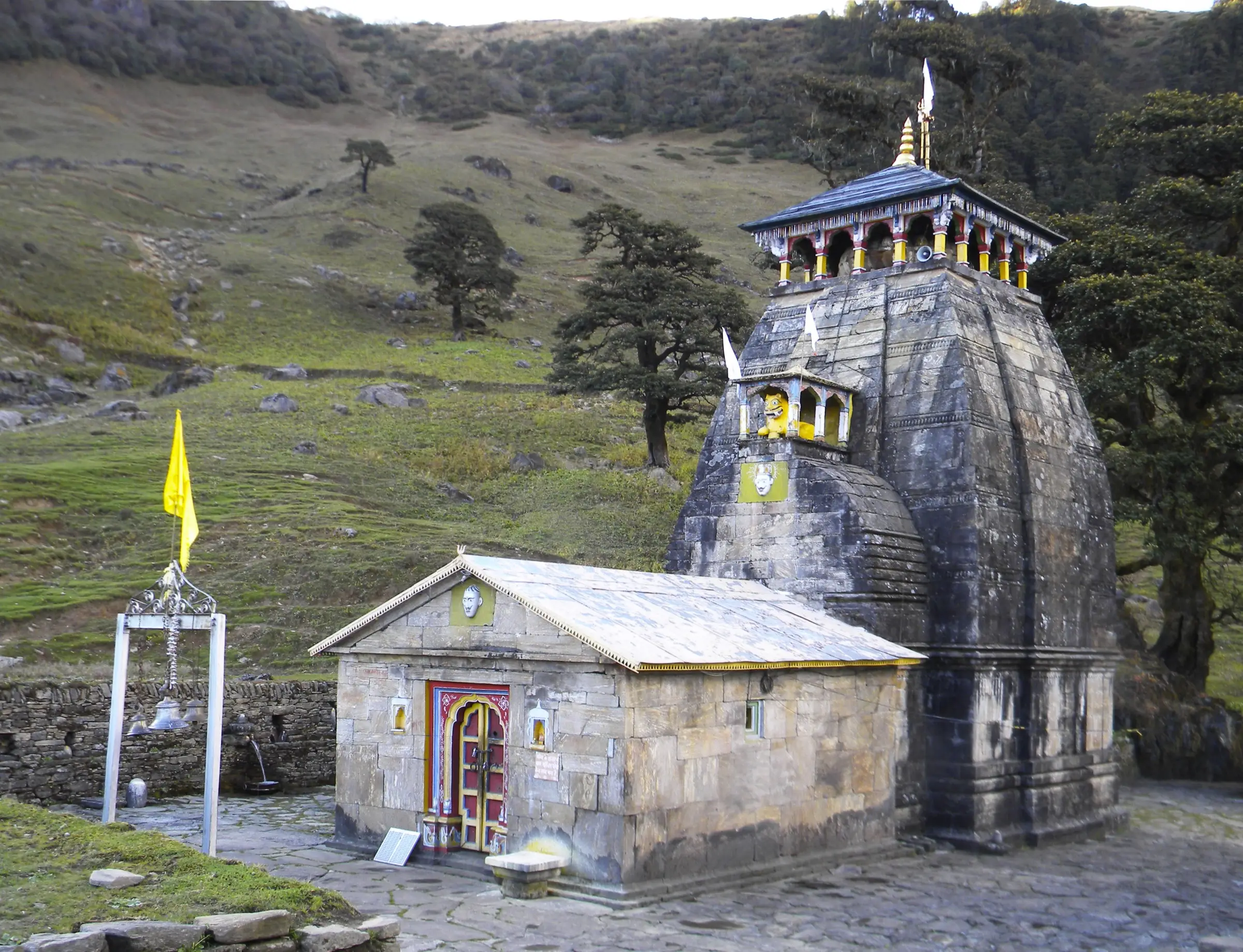 Madhyamaheshwar Temple, a dark stone Nagara-style shrine with a yellow flag, on a green meadow edged by trees