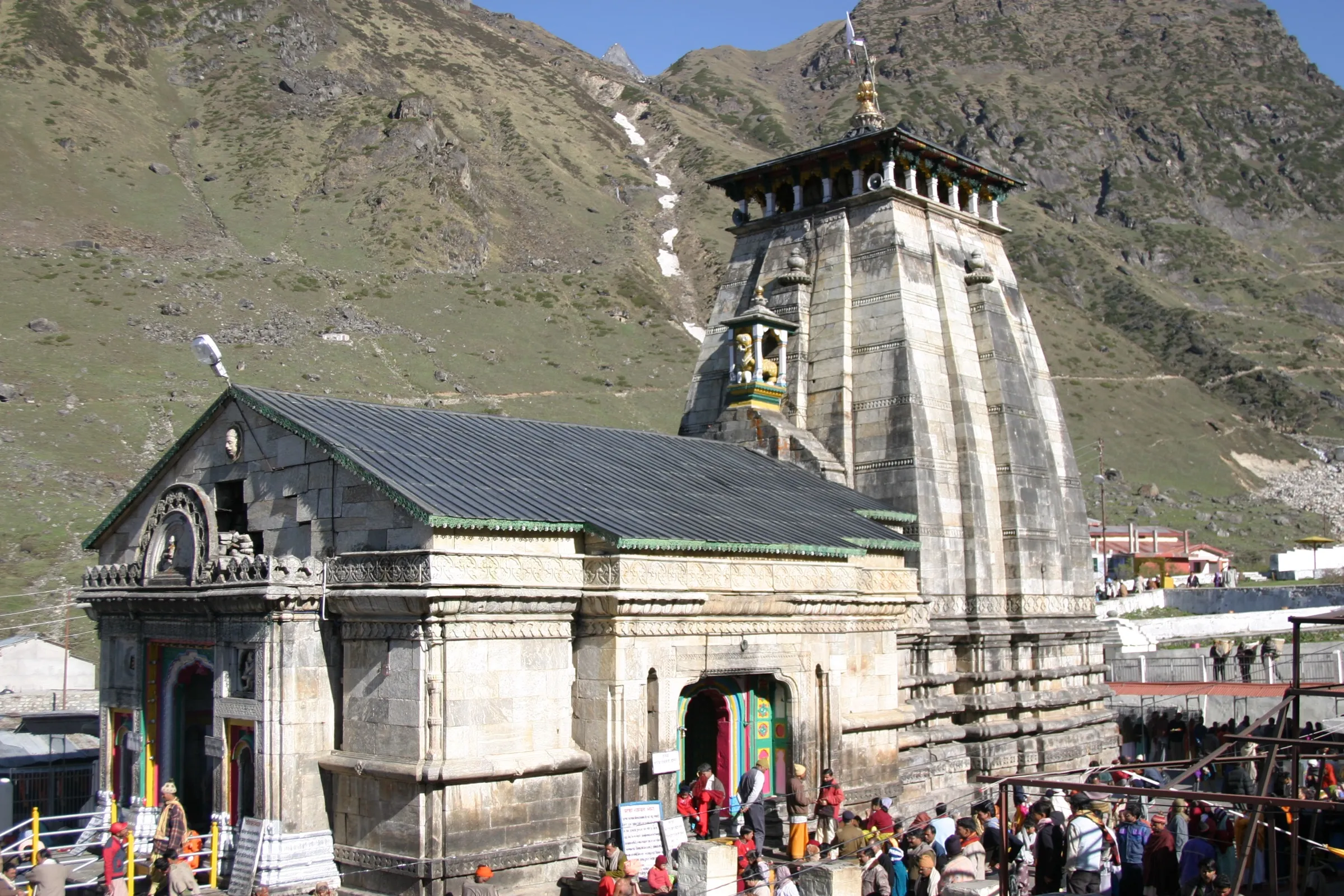 The grey stone Kedarnath temple with its tall shikhara and flag under green mountain slopes, with pilgrims in front of the doorway