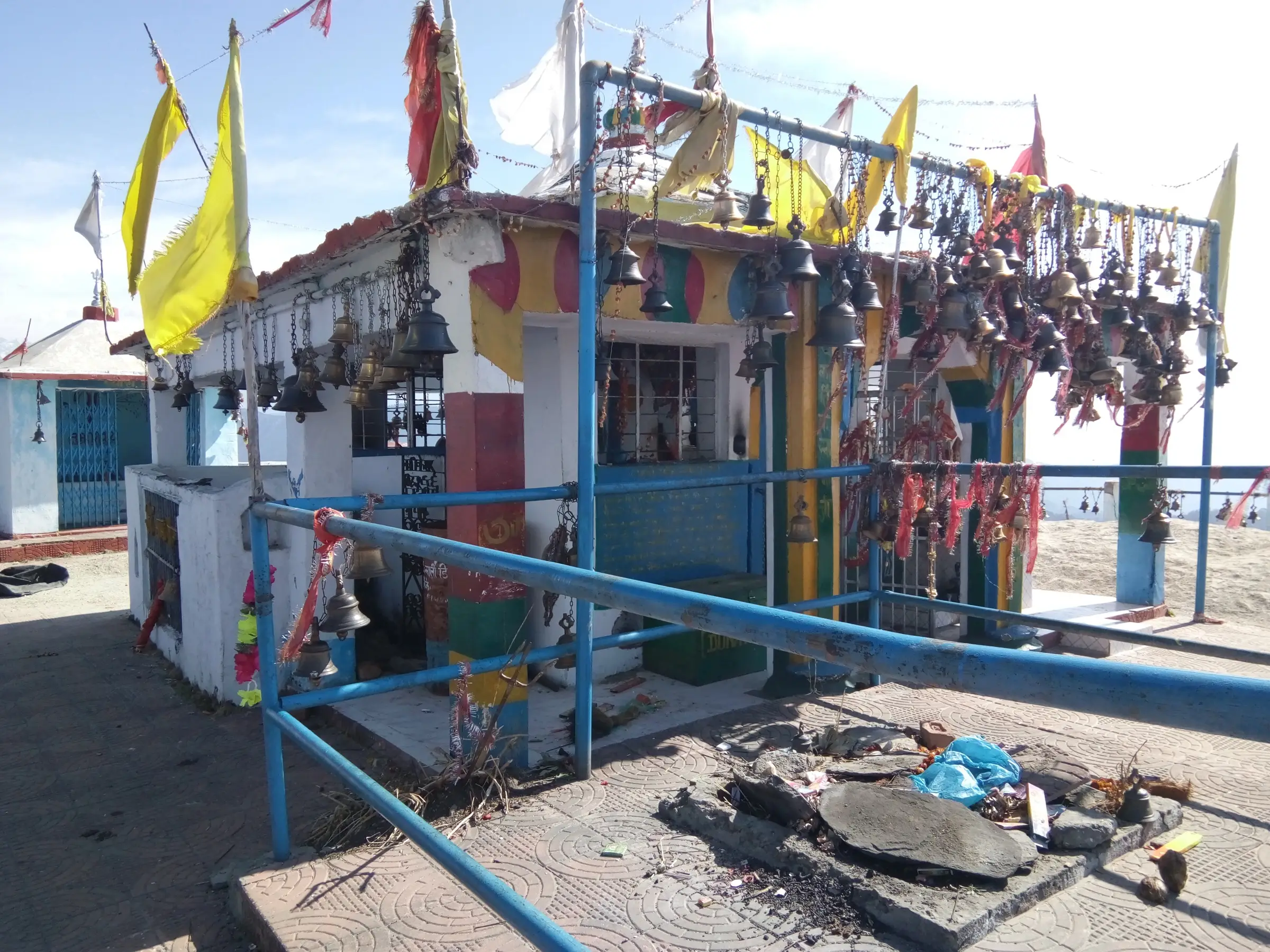 Colourful Kartik Swami shrine hung with hundreds of bells, red chunris and yellow flags under a clear sky
