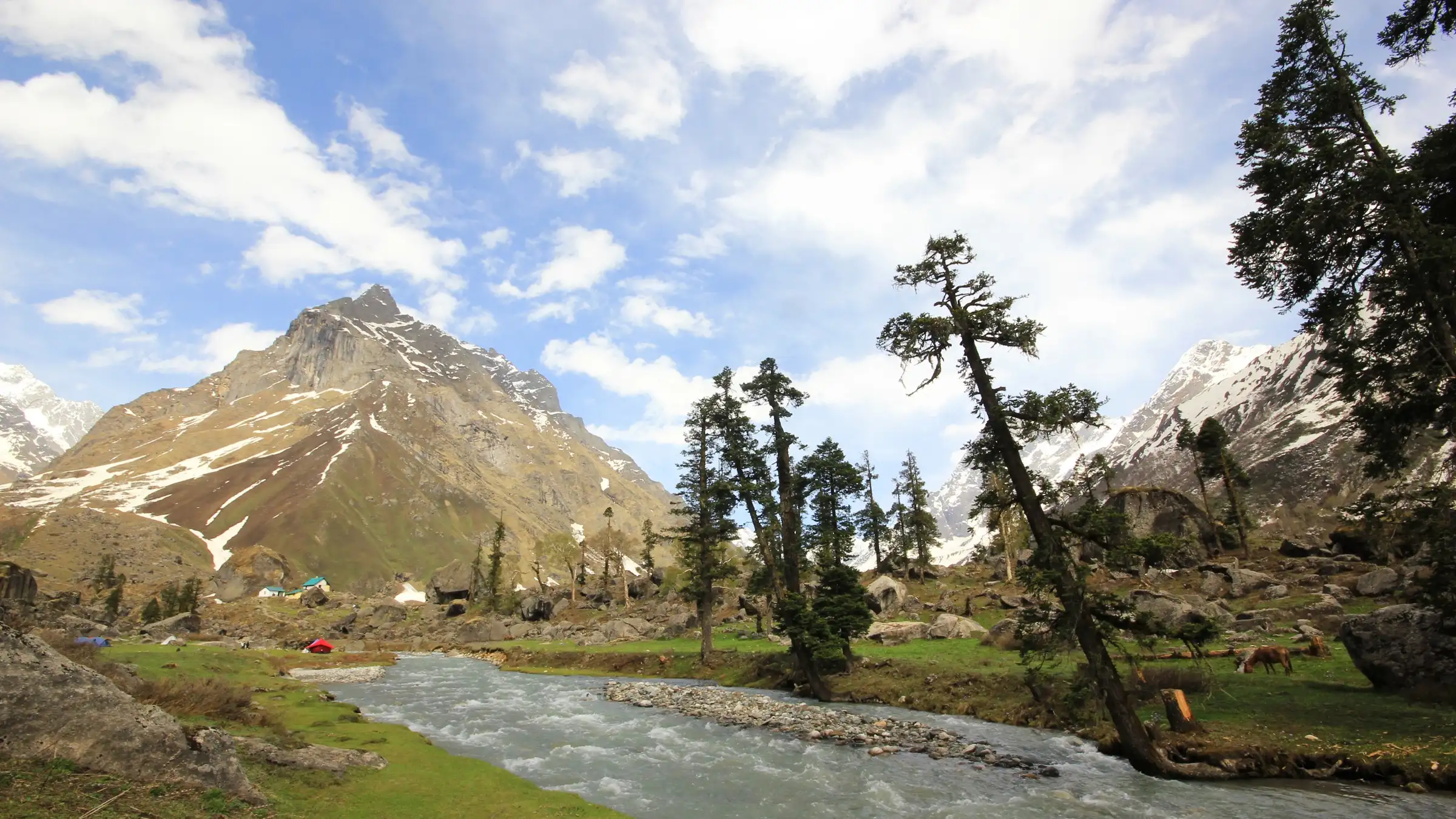 Tents and grazing horses on a meadow beside a rushing stream below a rocky snow-streaked peak in Har Ki Dun