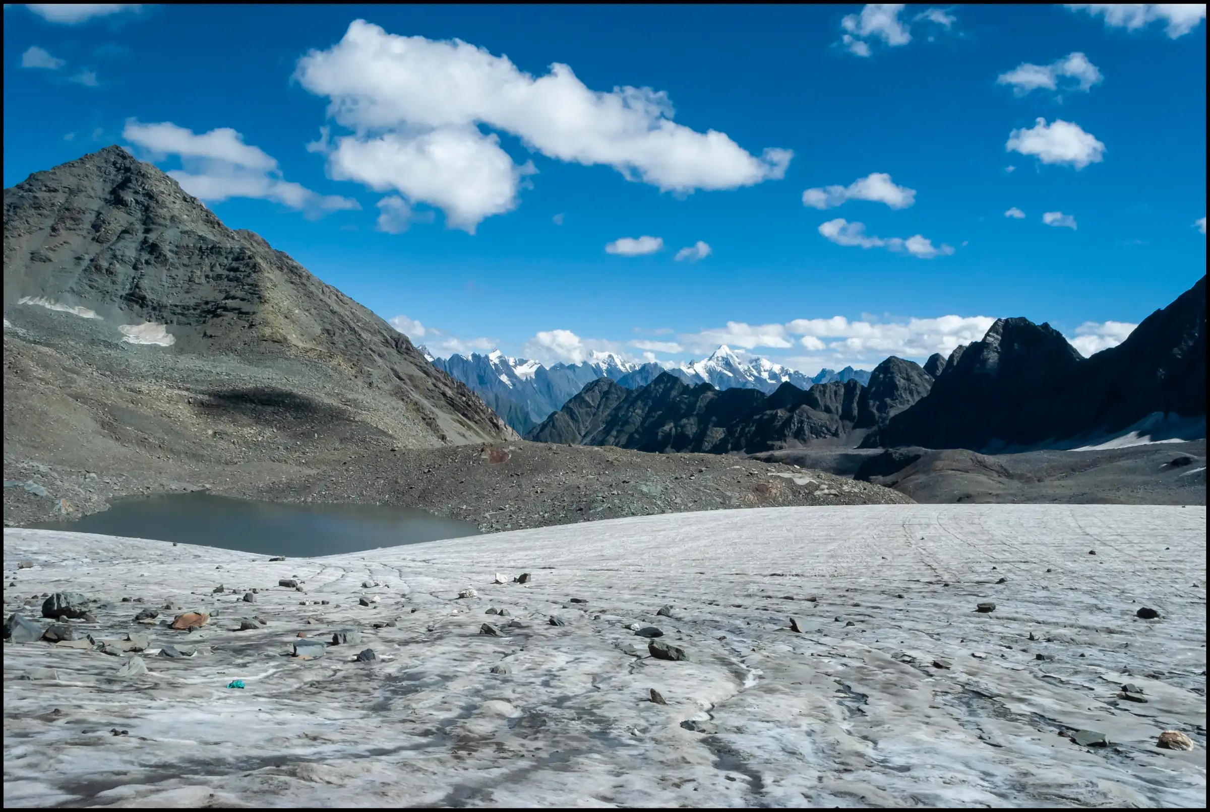 Grey glacier ice and moraine near Thamsar Pass with a small lake below dark rocky peaks and distant snow summits