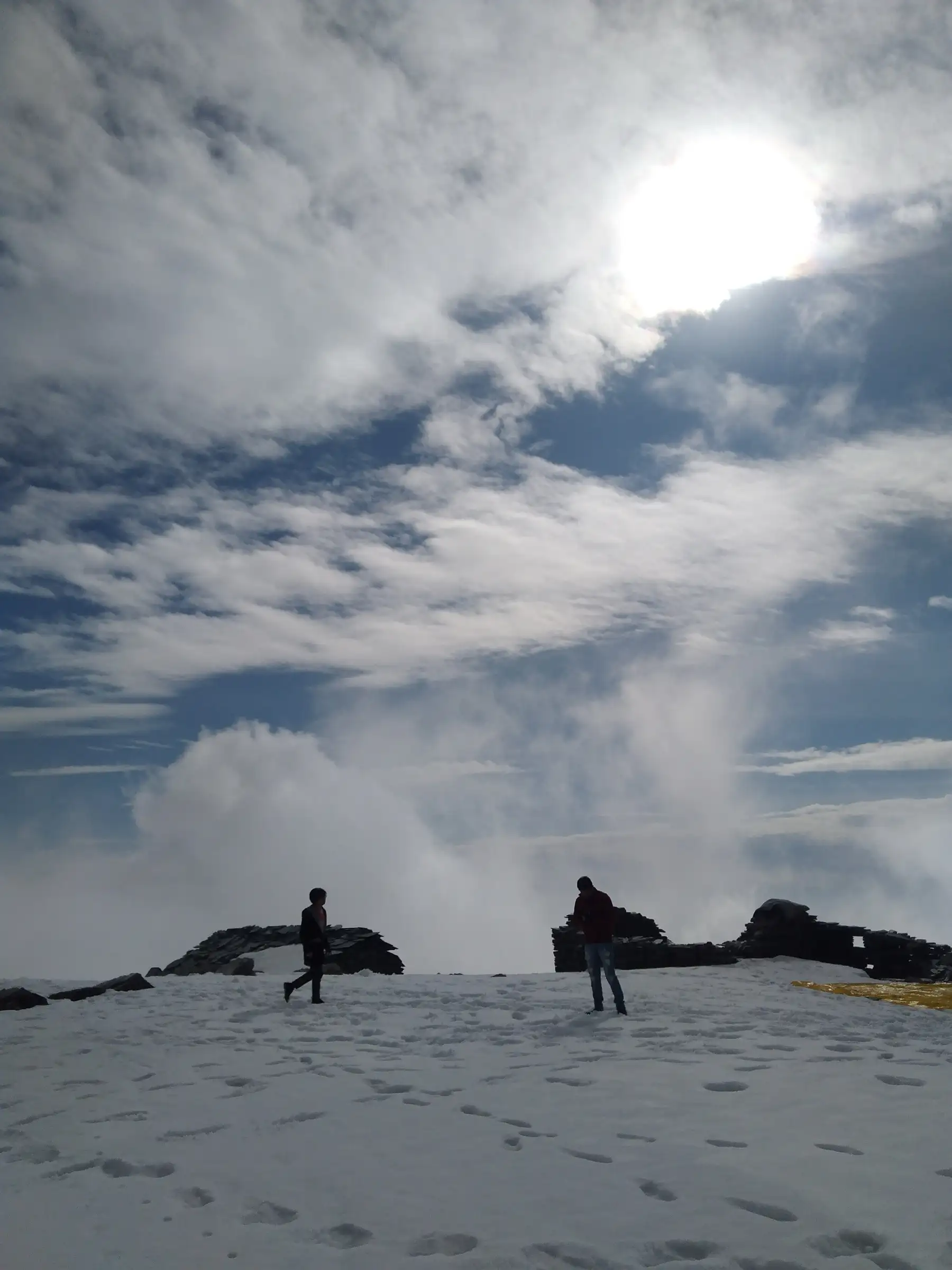 Two trekkers on the snow-covered Thatharana top in December, with stone huts and rising cloud