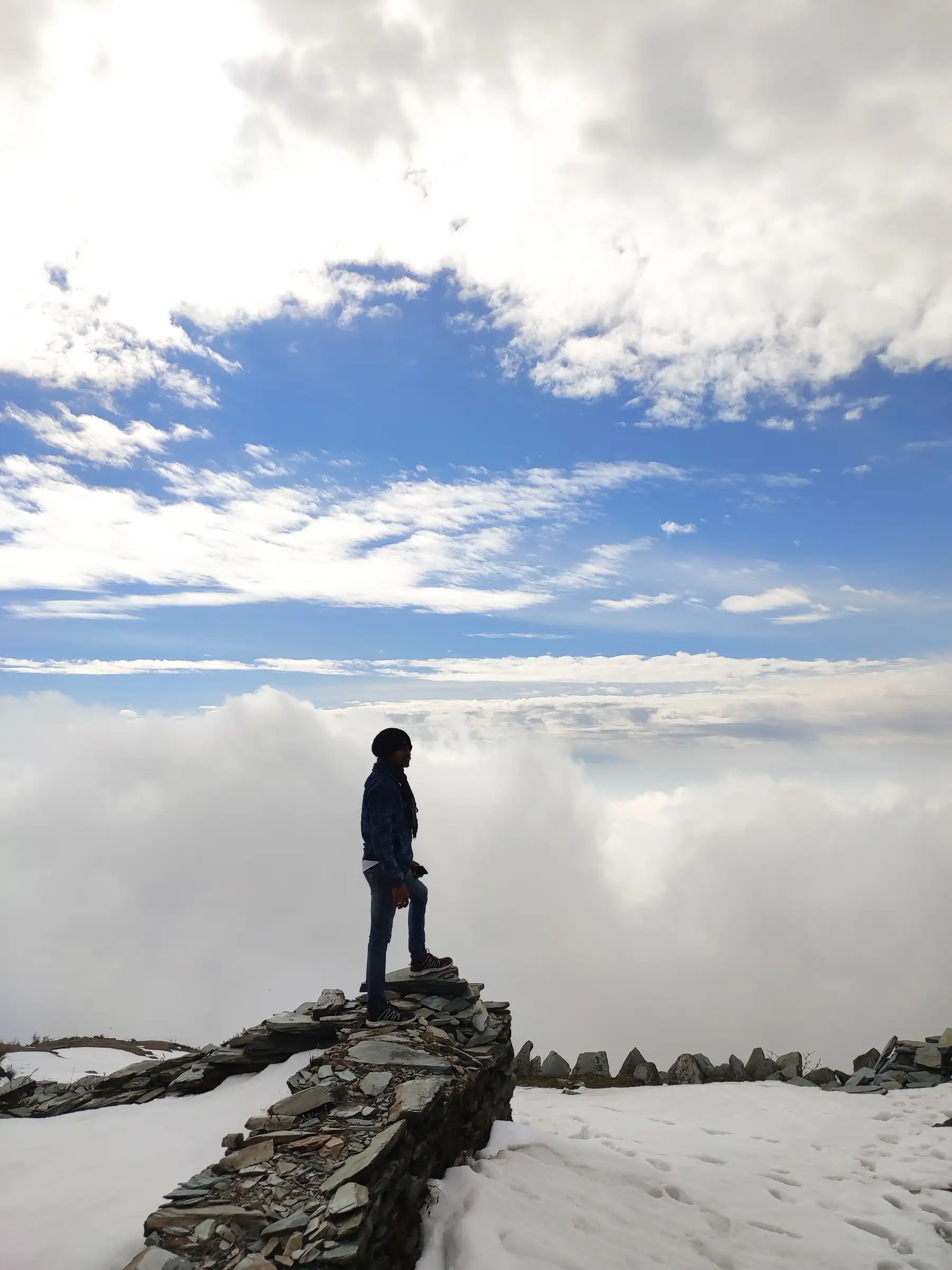 A trekker standing on a dry-stone wall in the snow at Thatharana, above a sea of cloud