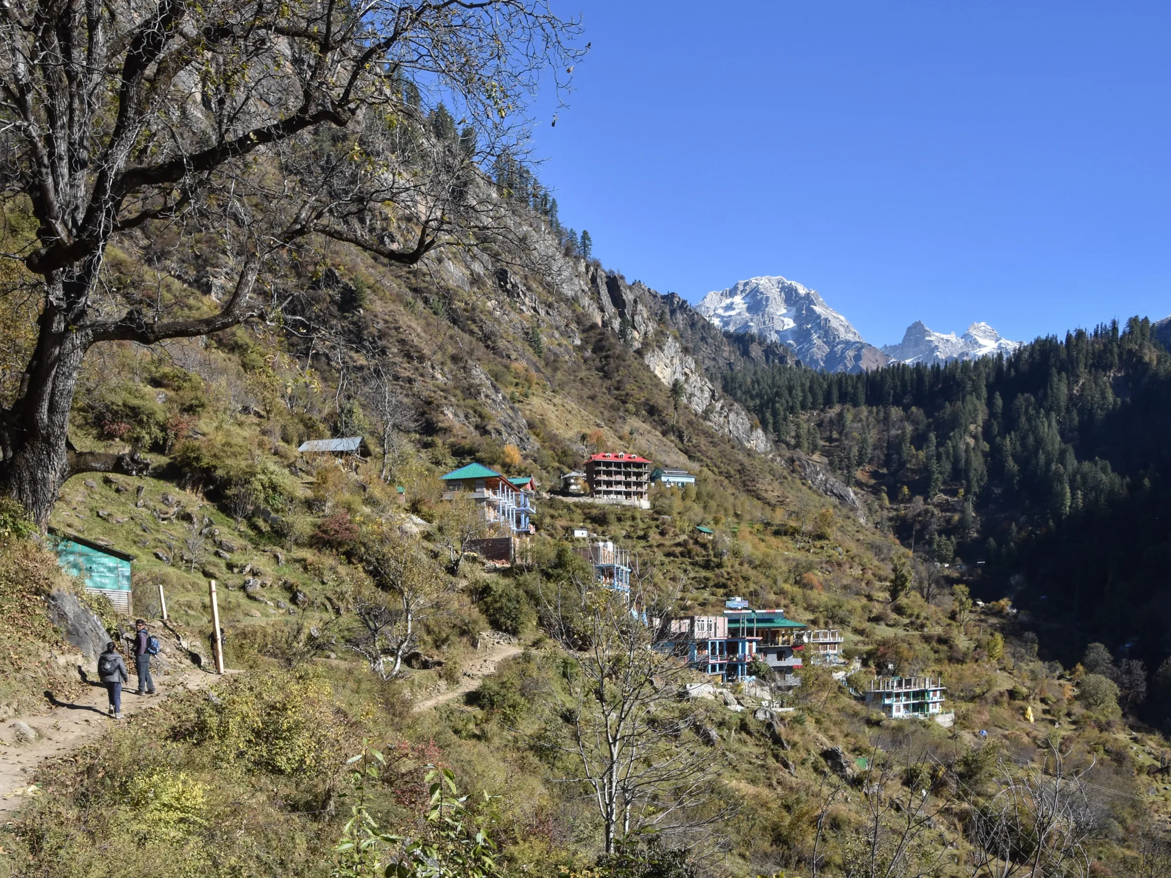 Two trekkers on the dirt trail out of Tosh towards Kutla, the start of the walk up the Tosh valley, with guesthouses on the slope and snow peaks behind