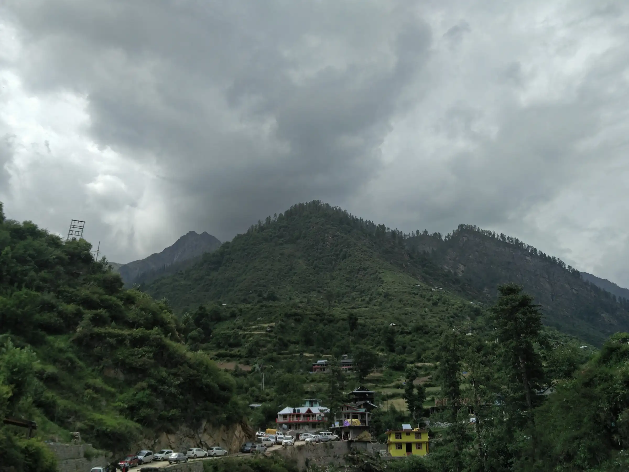 Road head and parked cars below Tosh, under forested hills and monsoon clouds, where the walk to the village begins