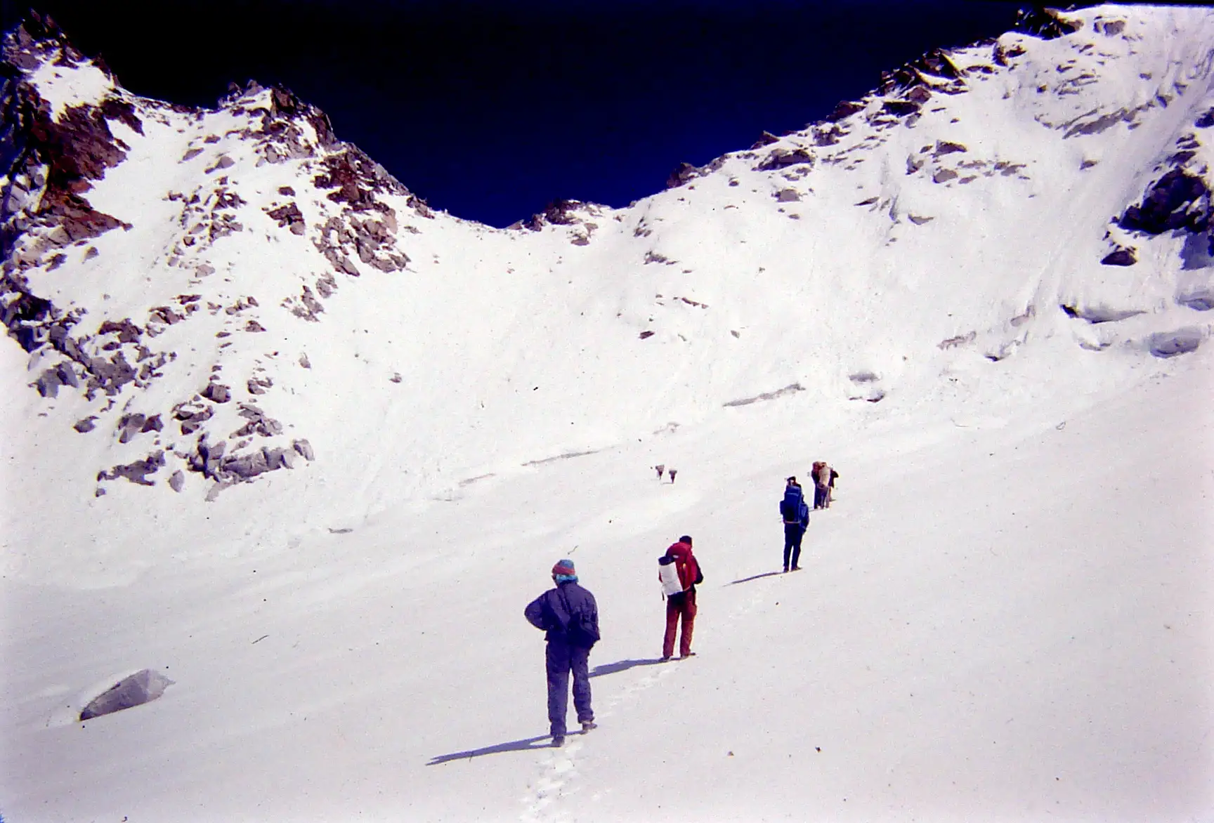 A line of trekkers walking up a snowfield towards the notch of Borasu Pass under a deep blue sky