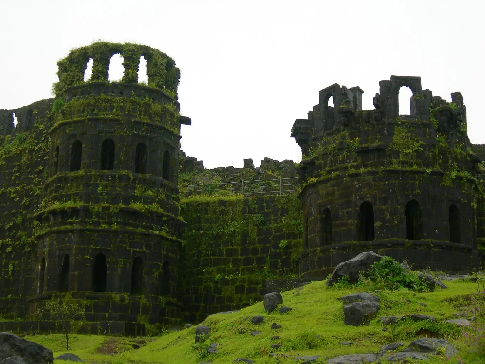 Two ruined round stone watchtowers (manore) of Raigad Fort, overgrown with moss above a grassy slope