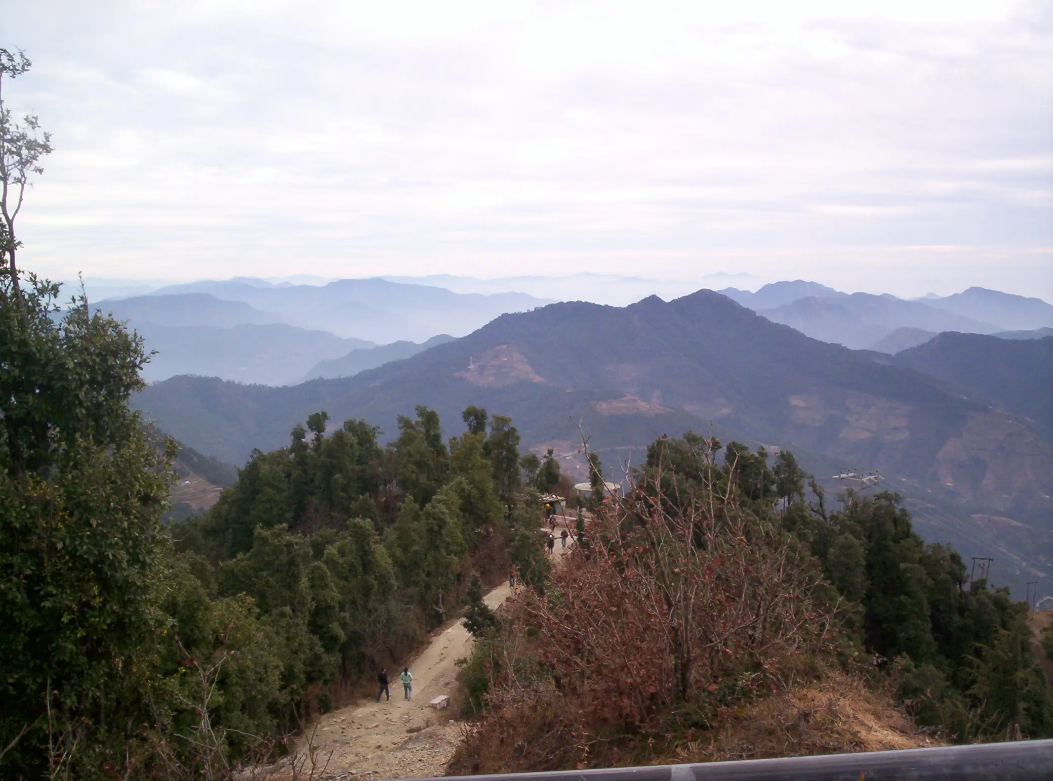 Footpath climbing a forested ridge towards Surkanda Devi with layered blue mountain ranges behind