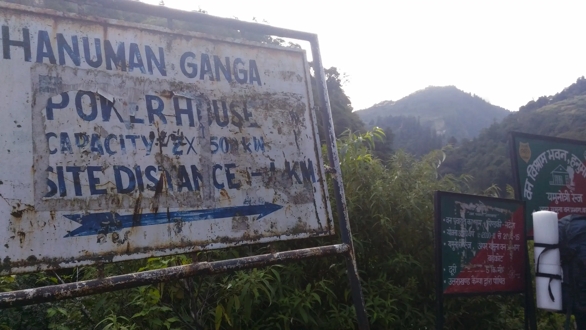 Faded Hanuman Ganga power house signboard and forest department boards beside the trail above Hanuman Chatti, with forested ridges behind